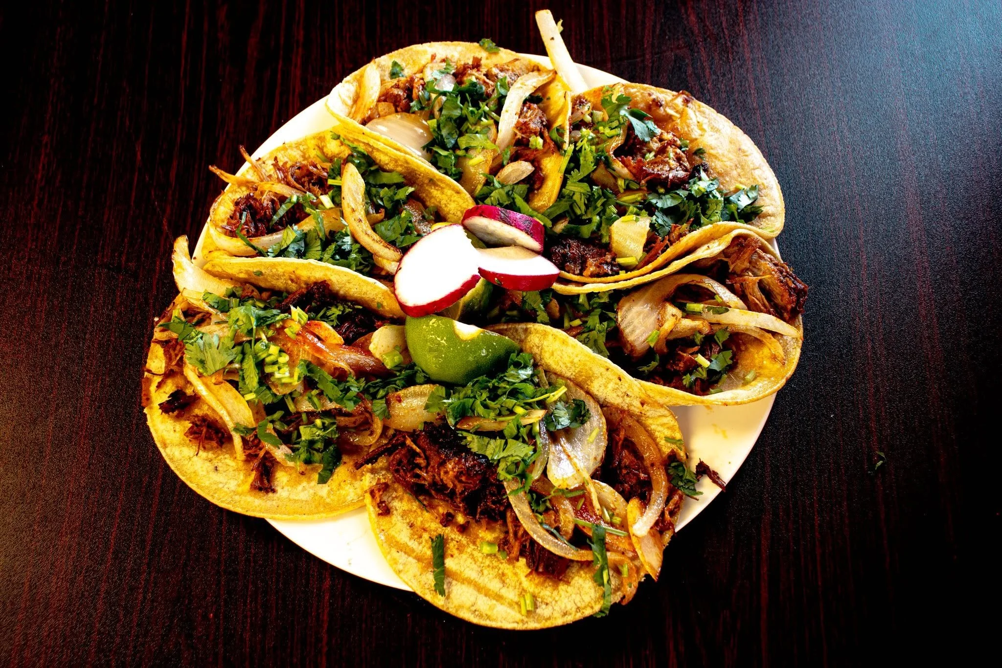 Three tacos filled with shredded meat, chopped herbs, onions, and sliced radishes on a white plate, placed on a dark wooden surface.