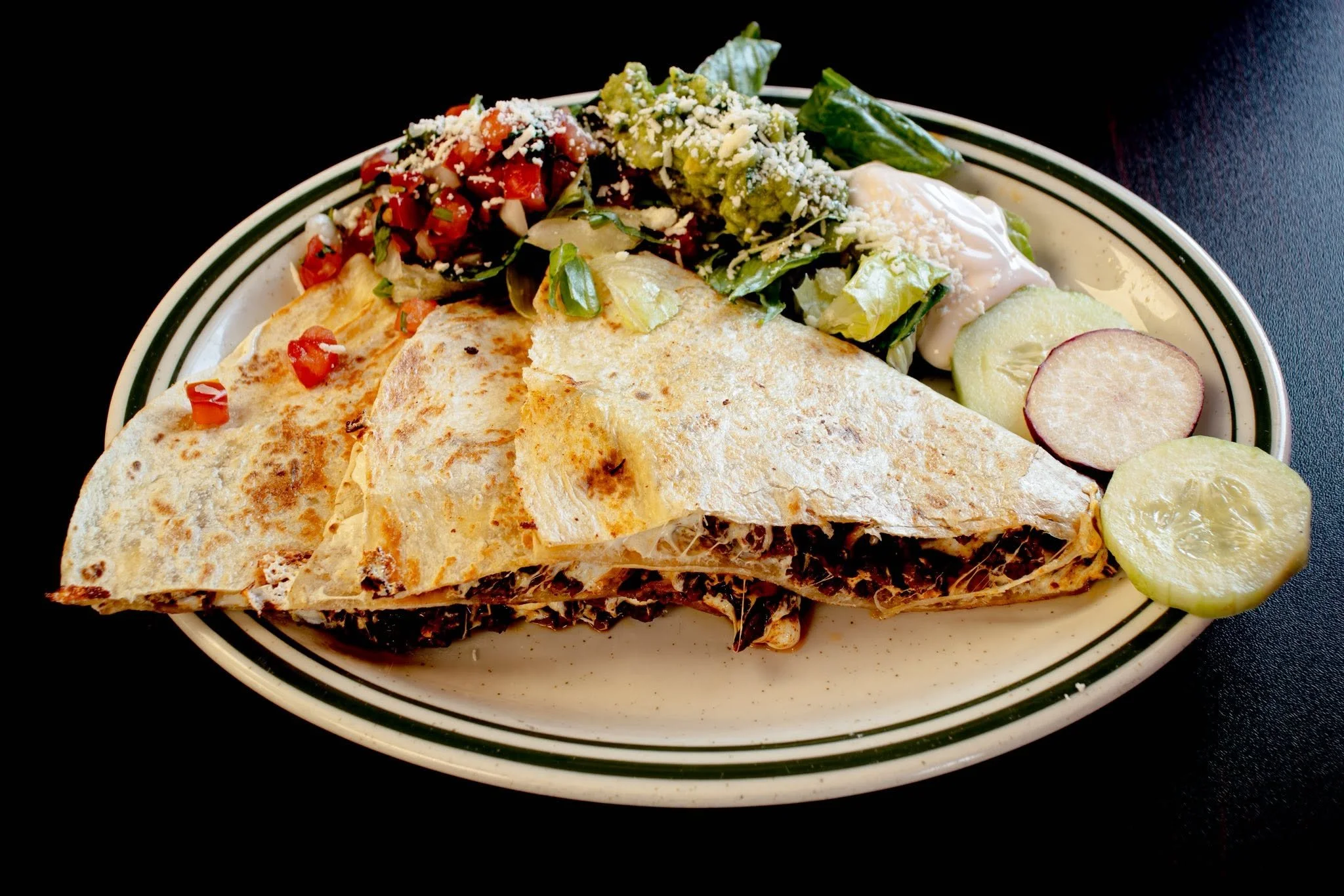 Plate with grilled quesadilla, fresh salsa, guacamole, and sliced limes on black background.