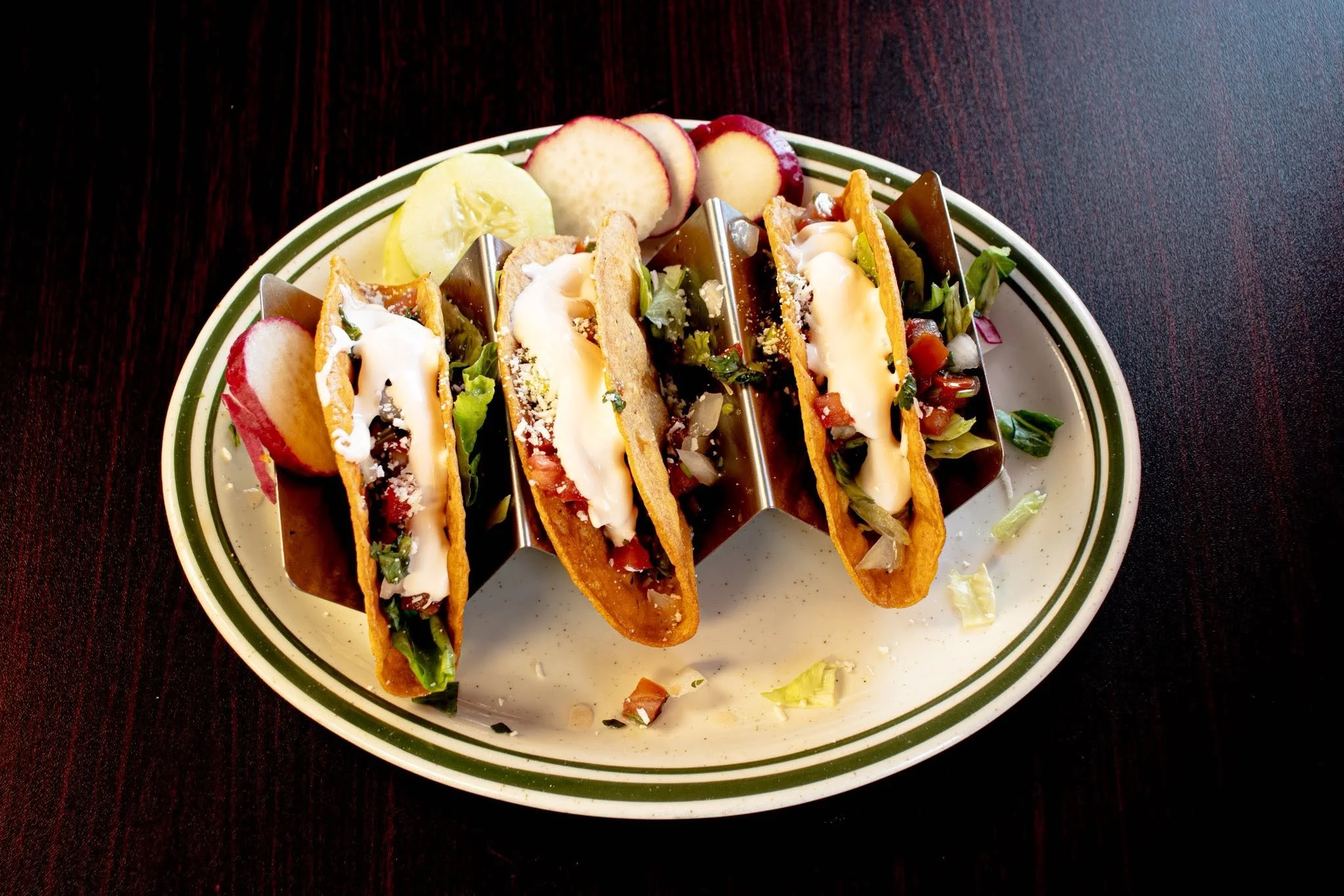 A plate of four beef tacos filled with lettuce, tomato, cheese, and sour cream, served with sliced cucumber, apple, and radish.