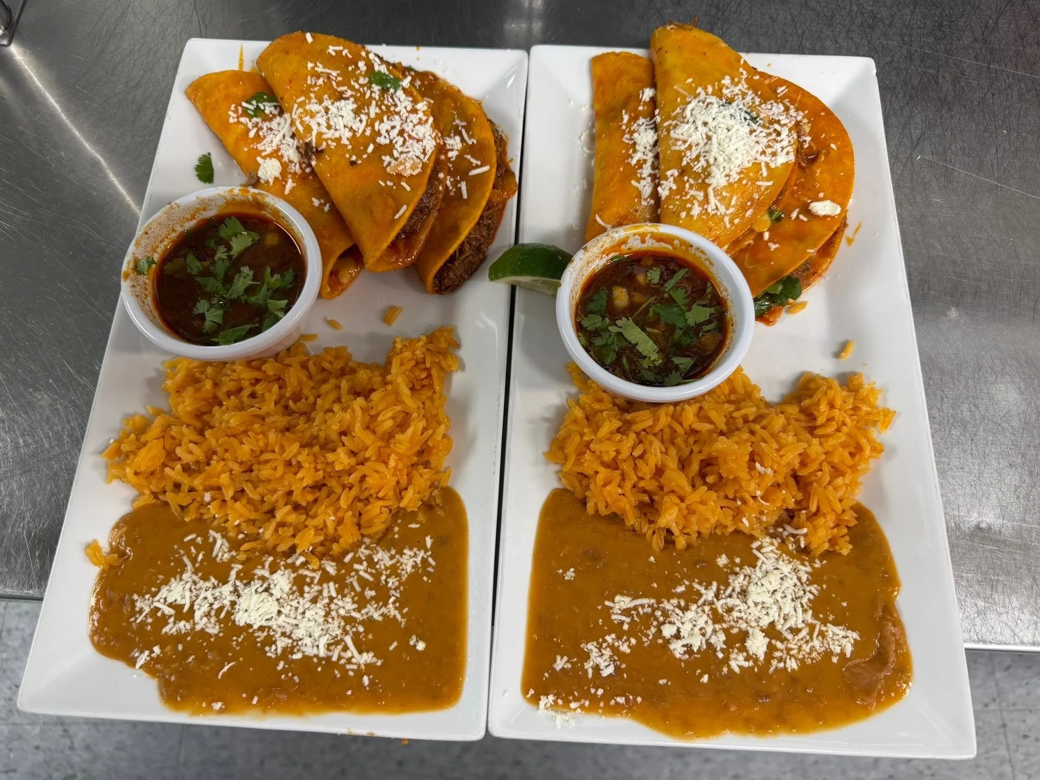 Two white rectangular plates with Mexican enchiladas, topped with shredded cheese and cilantro, served with rice and side bowls of dark enchilada sauce garnished with cilantro, set on a metal surface.