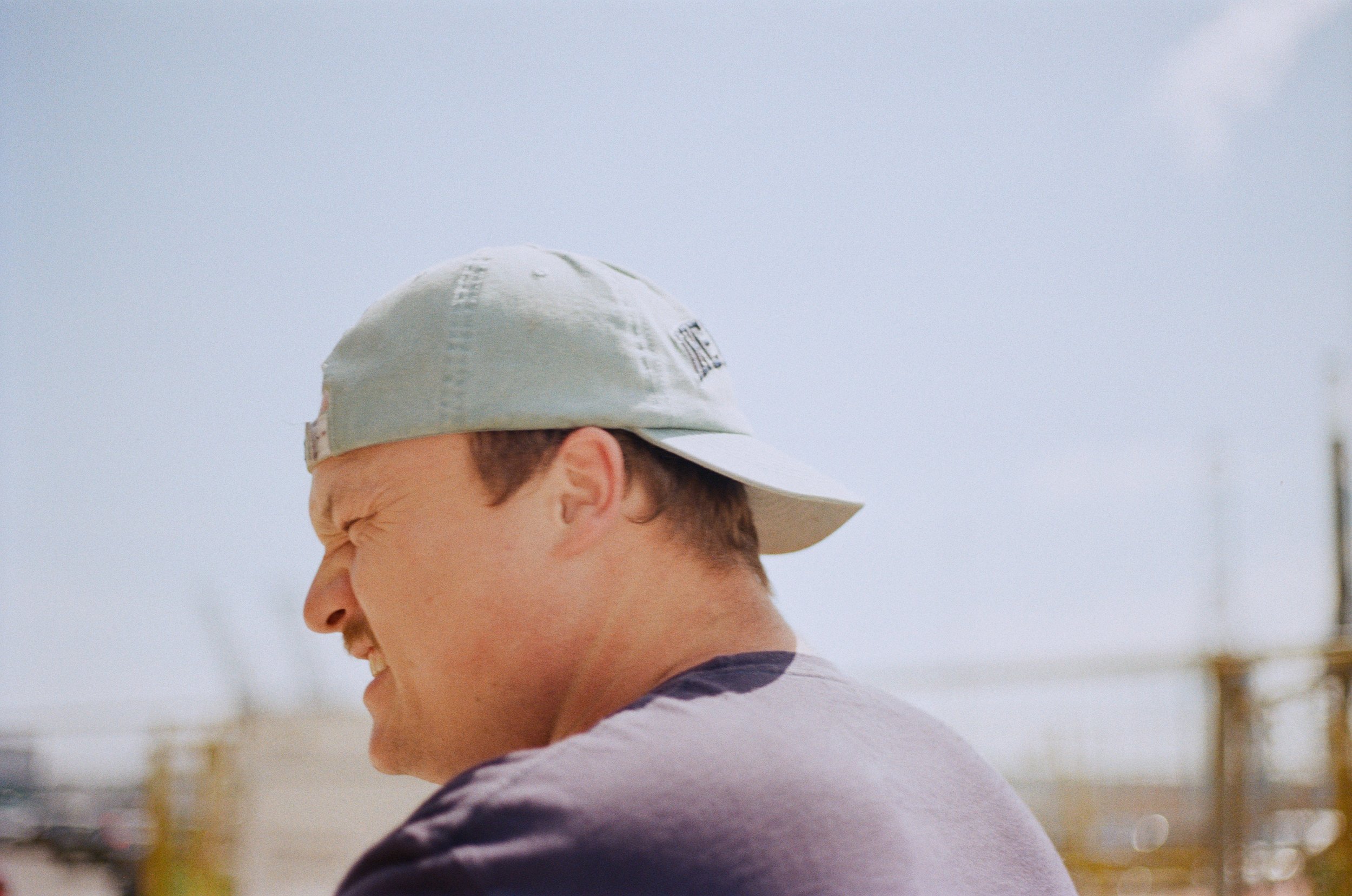 A person wearing a light-colored cap with a patch on the side, smiling and looking down, standing outdoors.