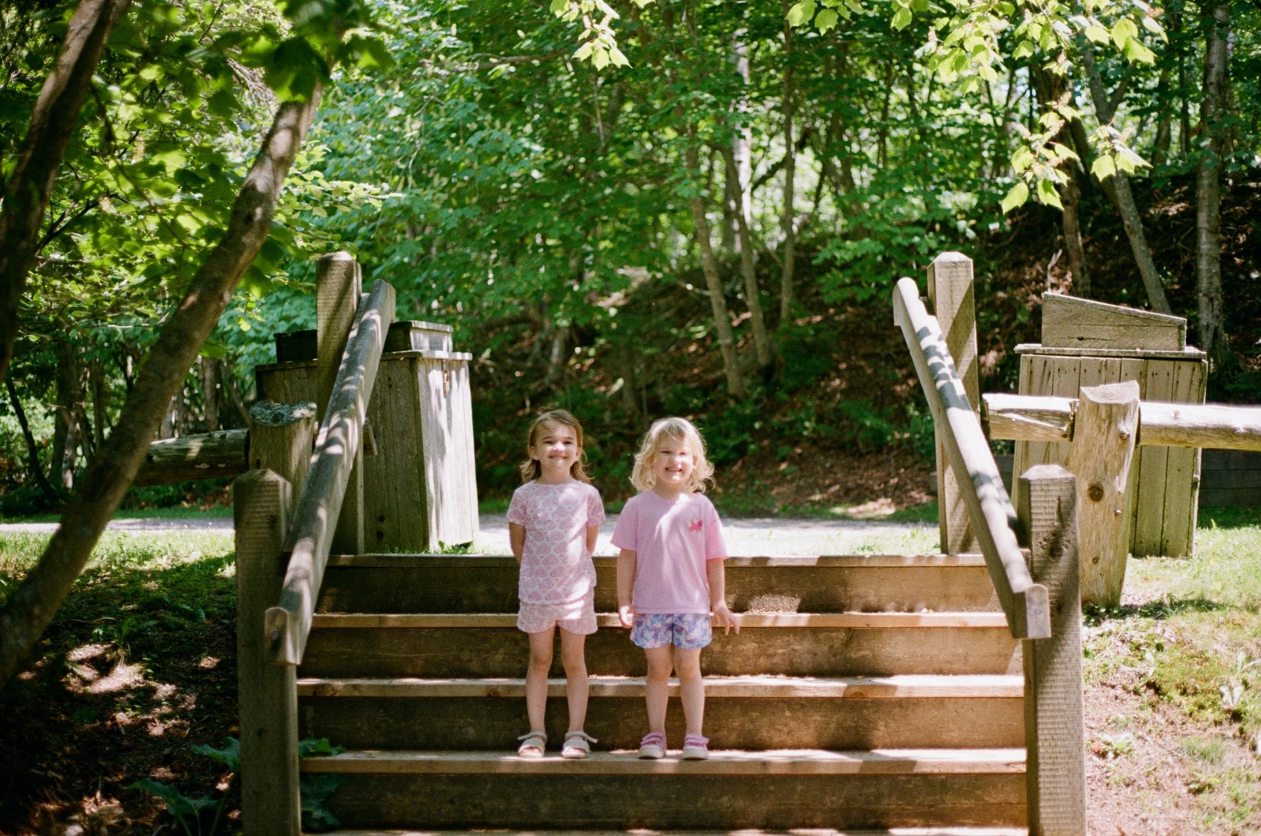 Two young girls standing on wooden steps at a playground, surrounded by green trees, smiling at the camera.