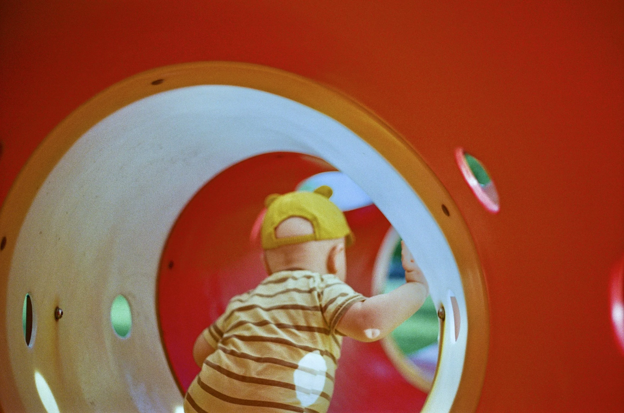 Child wearing a yellow cap and striped shirt playing inside a colorful tunnel at a playground.