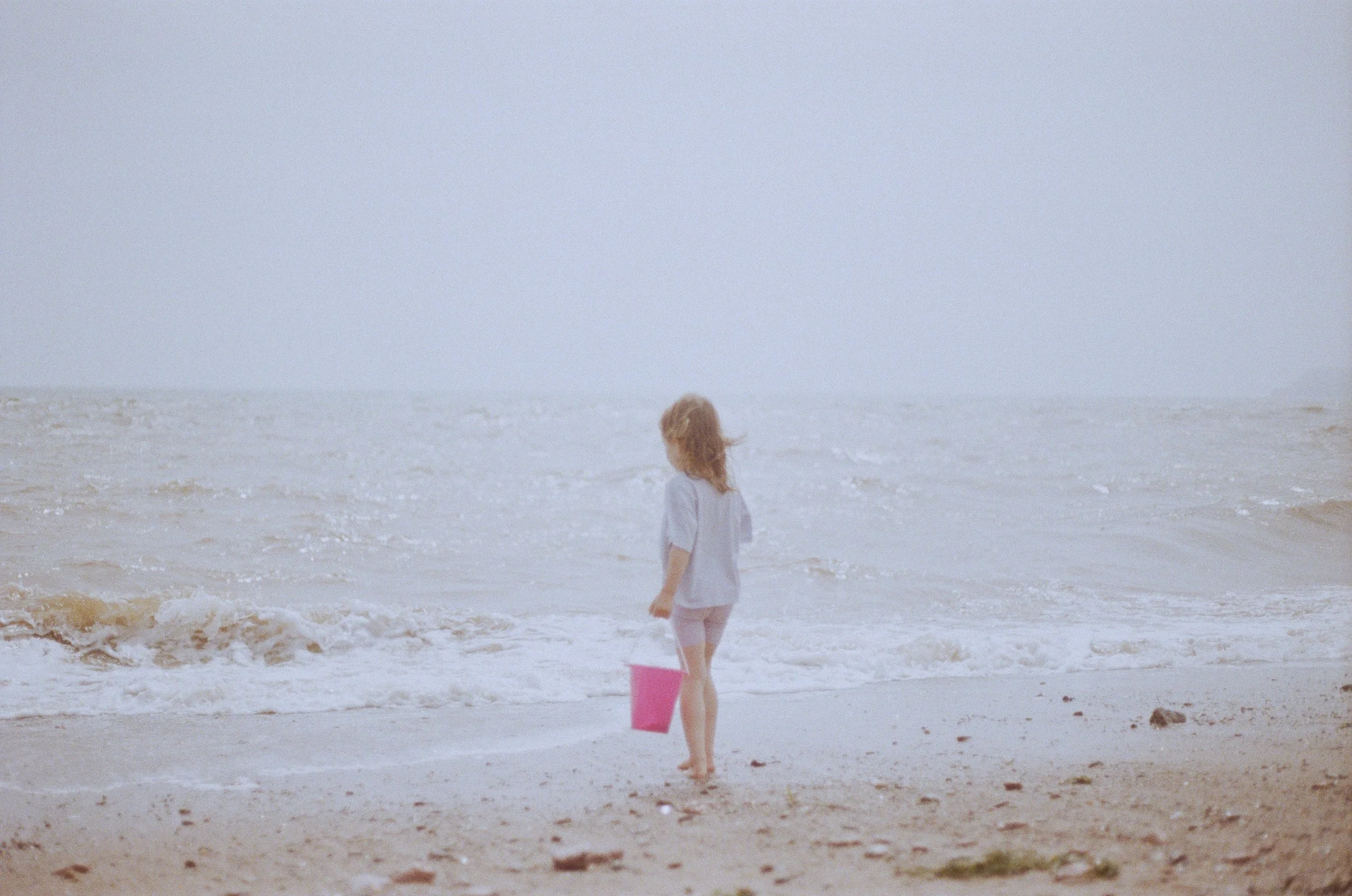 A young girl standing on the beach facing the ocean, holding a pink bucket.