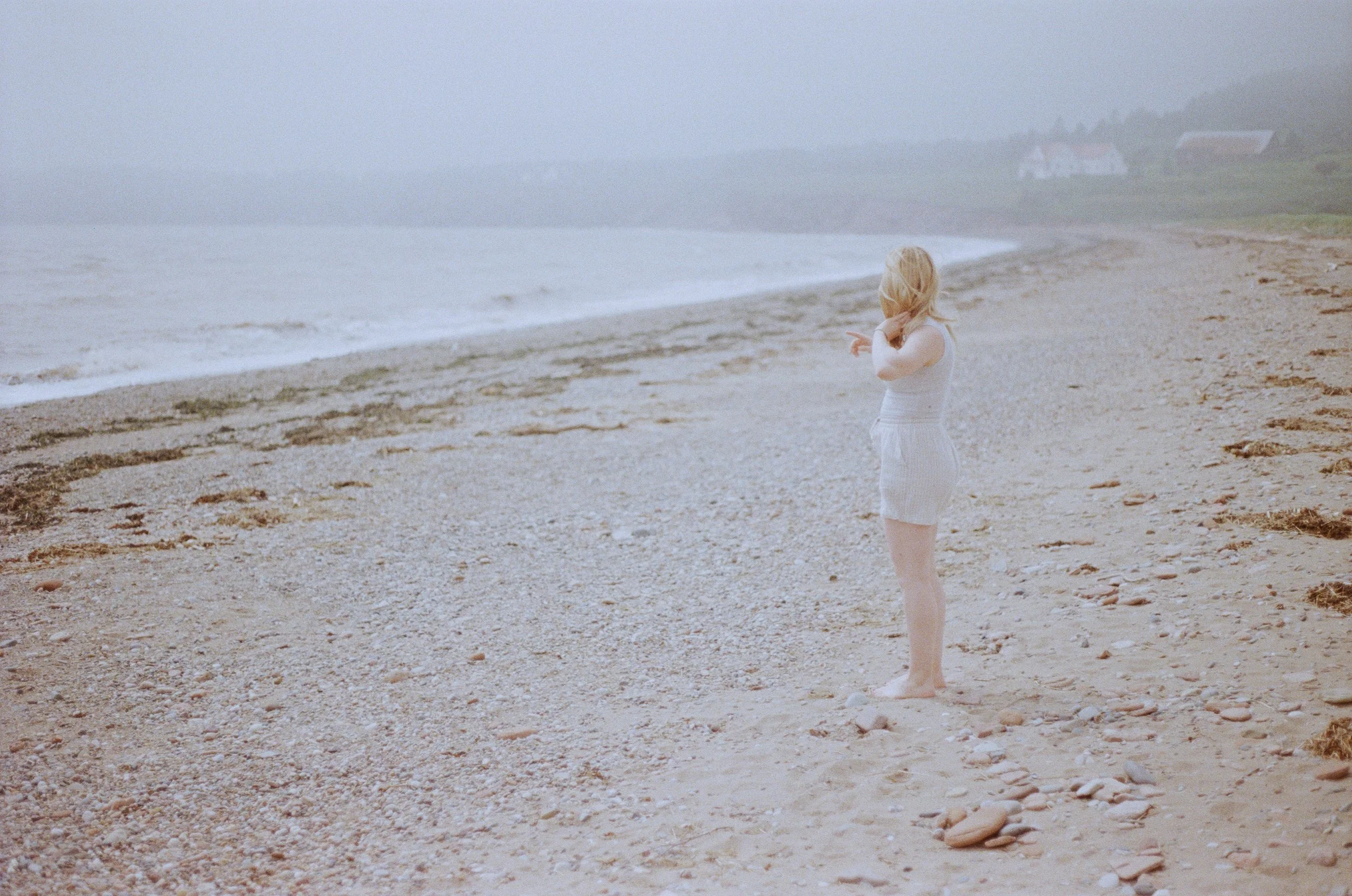 A woman with blonde hair standing barefoot on a beach, looking towards the ocean, with foggy weather and a few houses in the distance.