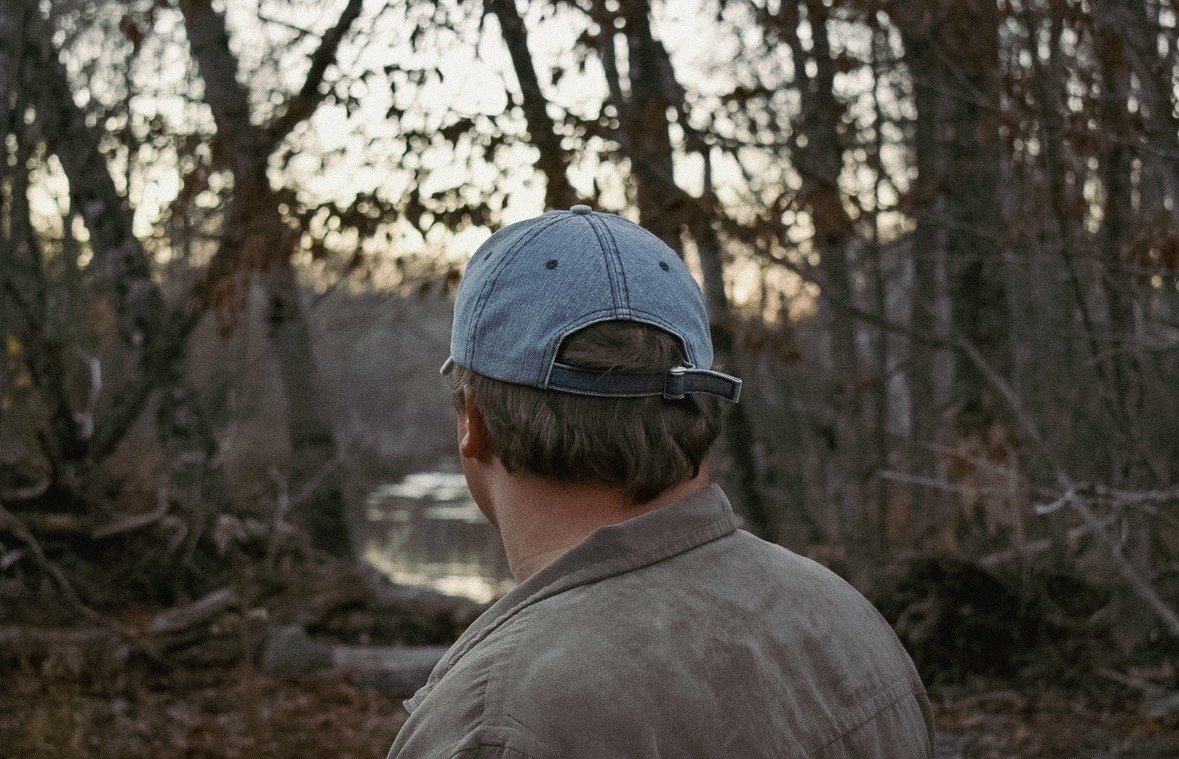 A man wearing a blue baseball cap and tan jacket stands outdoors, looking at a creek in a wooded area during sunset.