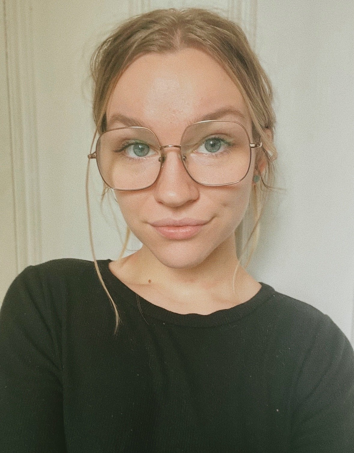 Close-up of a young woman with light brown hair, wearing glasses and a black shirt, smiling softly at the camera against a neutral background.