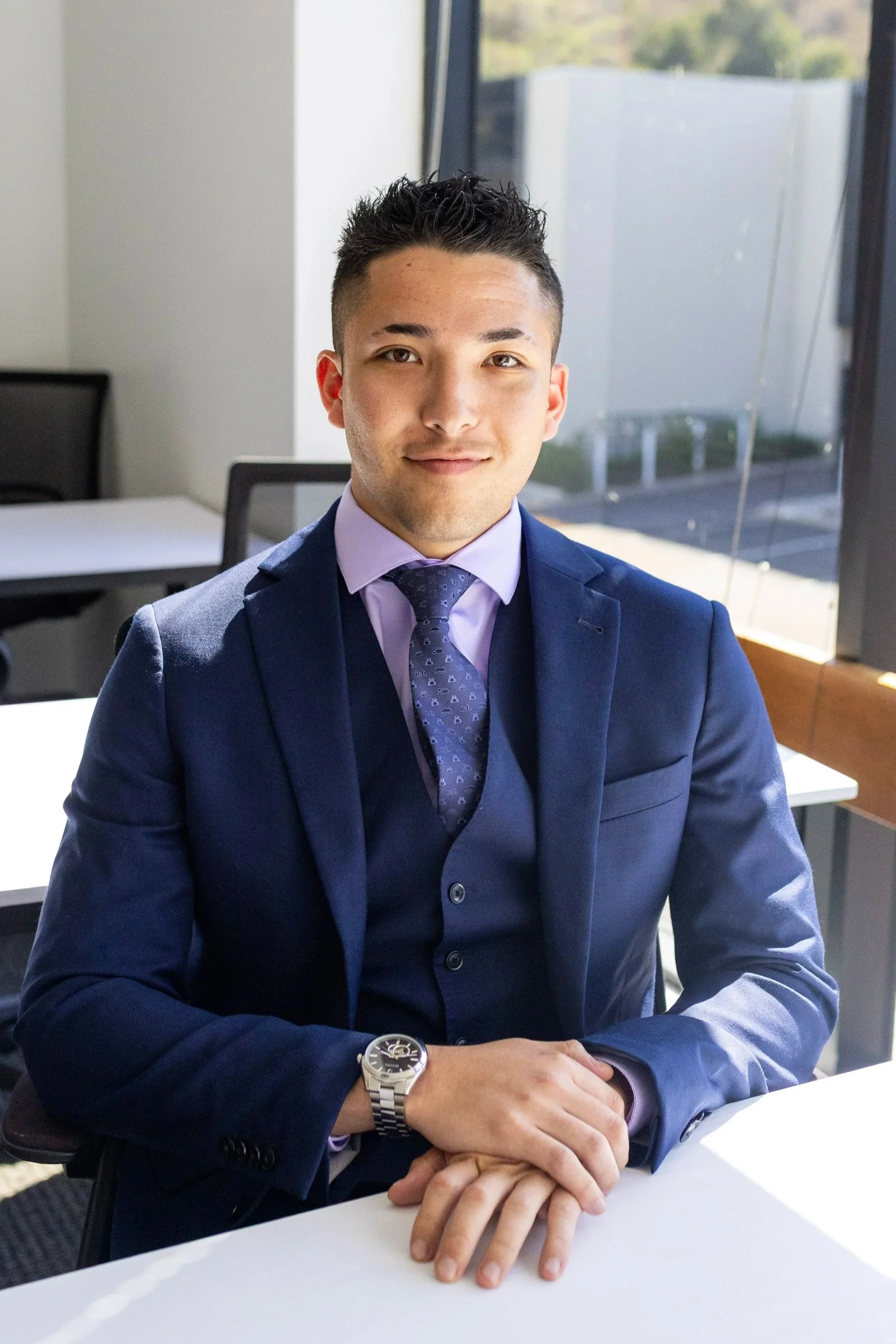 A young man wearing a navy suit and purple shirt sitting at a conference table in an office with large windows, smiling with hands resting on the table, wearing a silver watch.