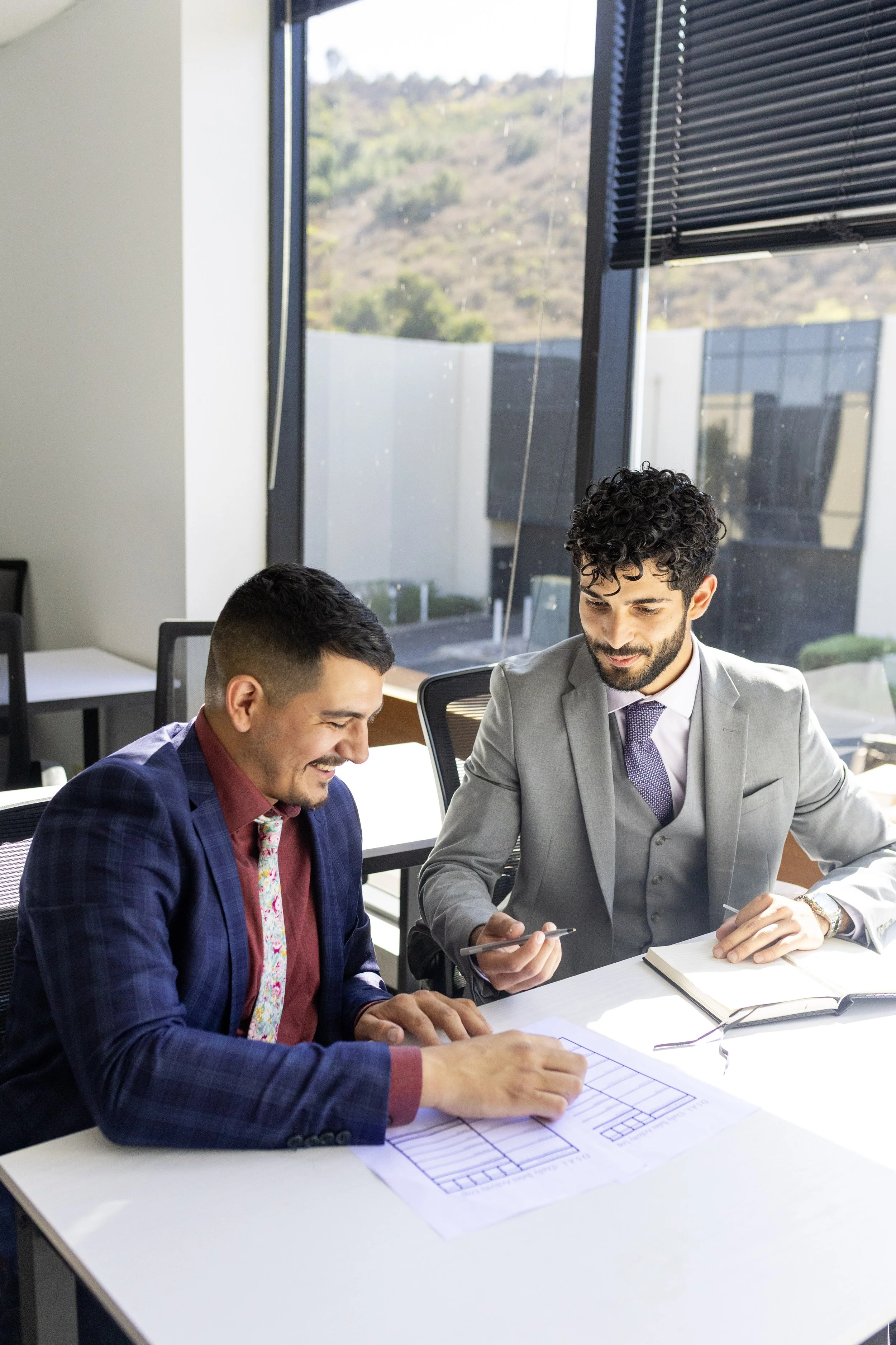 Two men in business attire sitting at a white table in a modern office during the daytime. They are reviewing documents with blueprints spread out on the table. One man, with a beard and curly hair, is in a gray suit and discussing with the other man, who has short hair and is wearing a blue checkered suit. Large windows with black blinds and a view of hills outside are in the background.