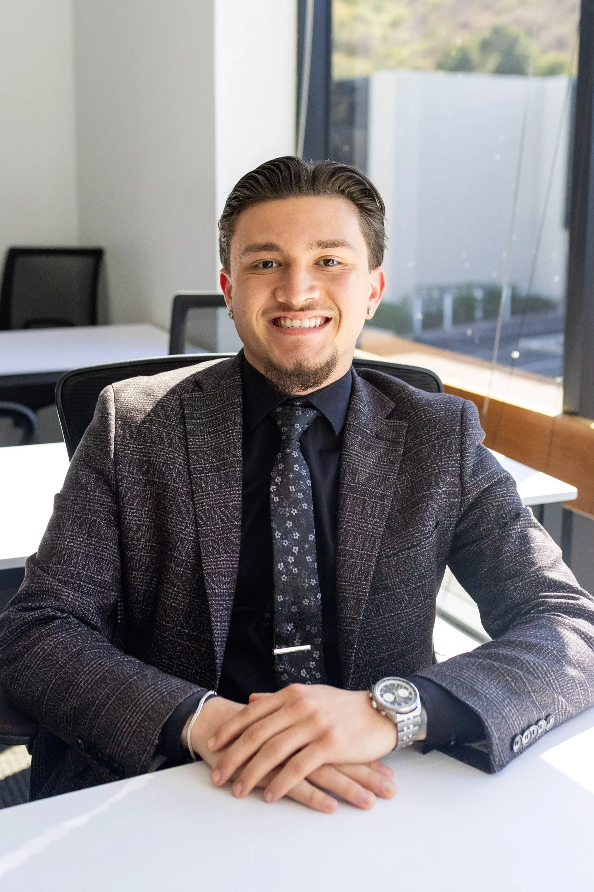 A young man in a suit sitting at a white table in an office, smiling at the camera, with large windows showing an outdoor view.