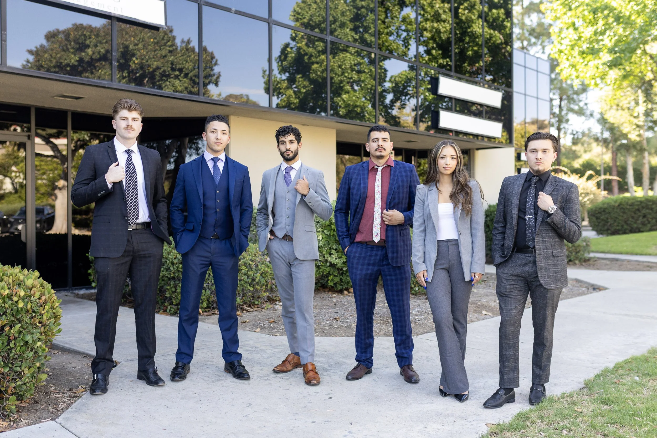 Group of six professionally dressed young adults standing outside in front of a modern office building with reflective glass windows, greenery, and trees.