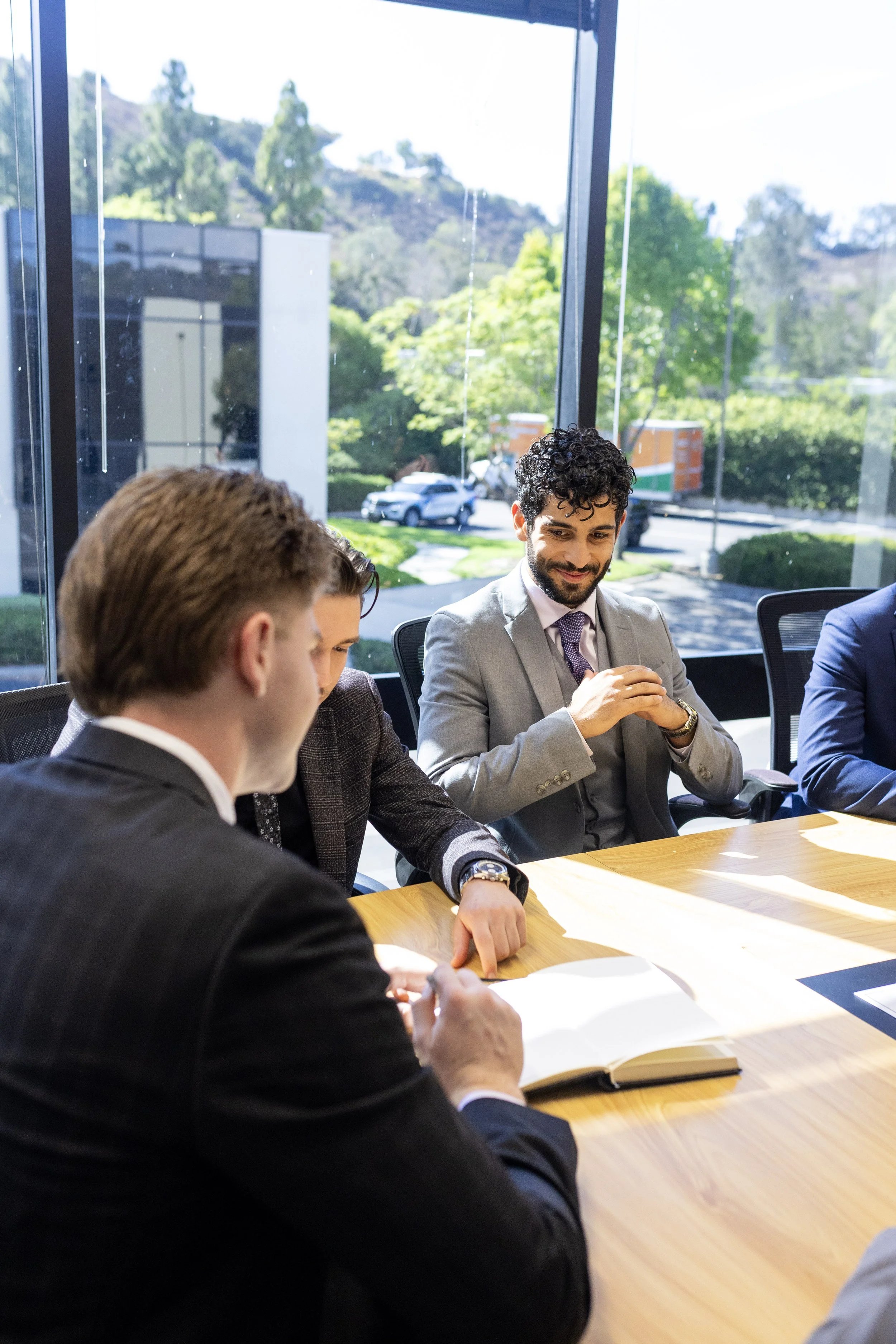 A group of business professionals sitting around a conference table during a meeting, with a view of the outdoors through large glass windows.