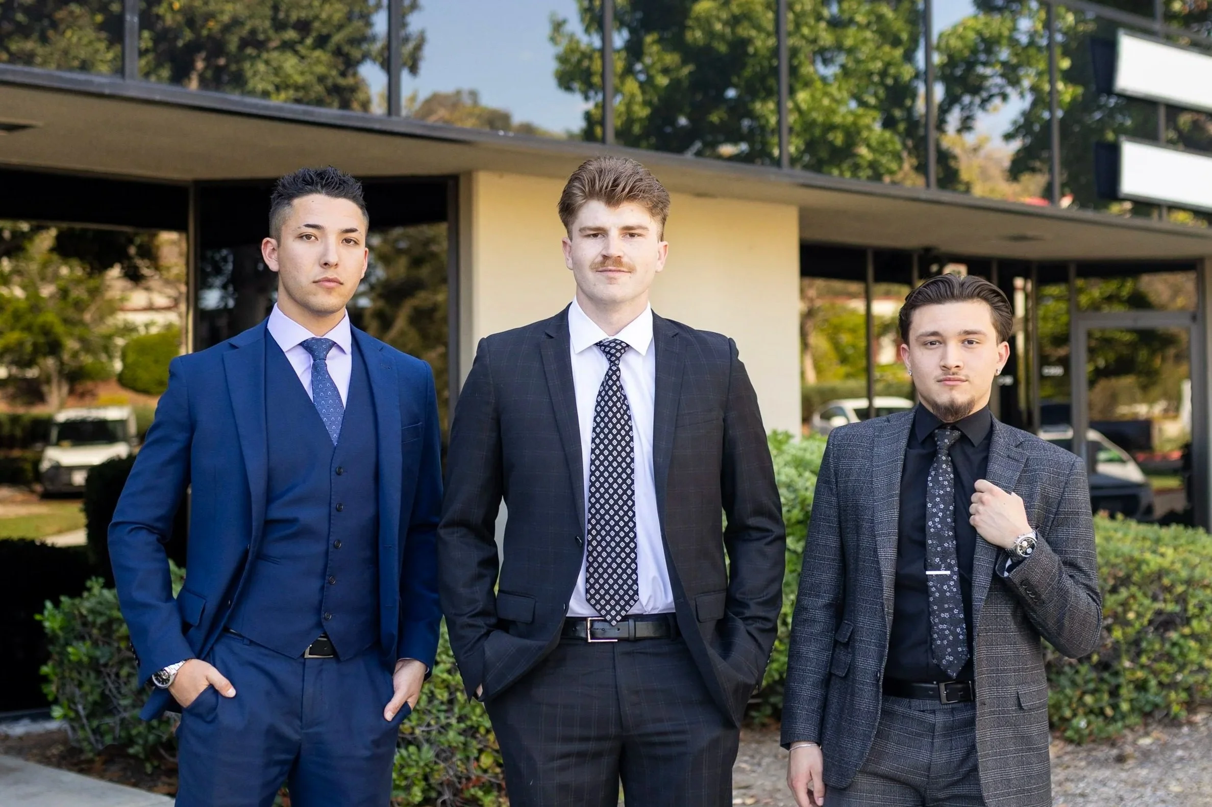 Three young men wearing suits standing outdoors in front of a modern building with green trees in the background.