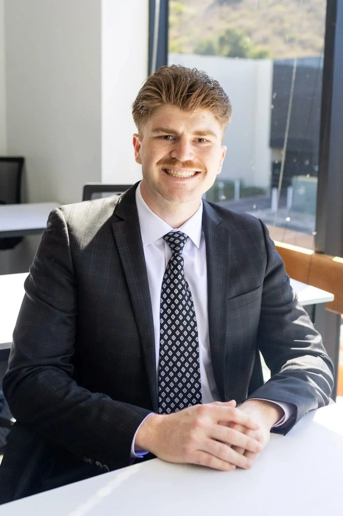 Portrait of a smiling man in a business suit sitting at a table in an office with large windows and a view of trees outside.