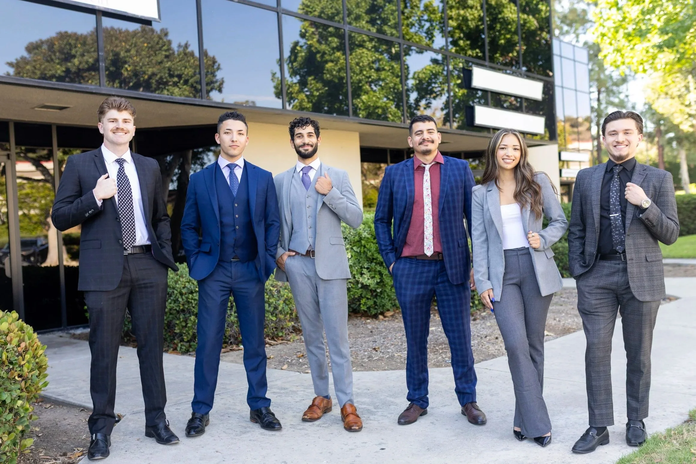 Group of six professionally dressed young adults standing outside in front of a glass building, smiling at the camera.