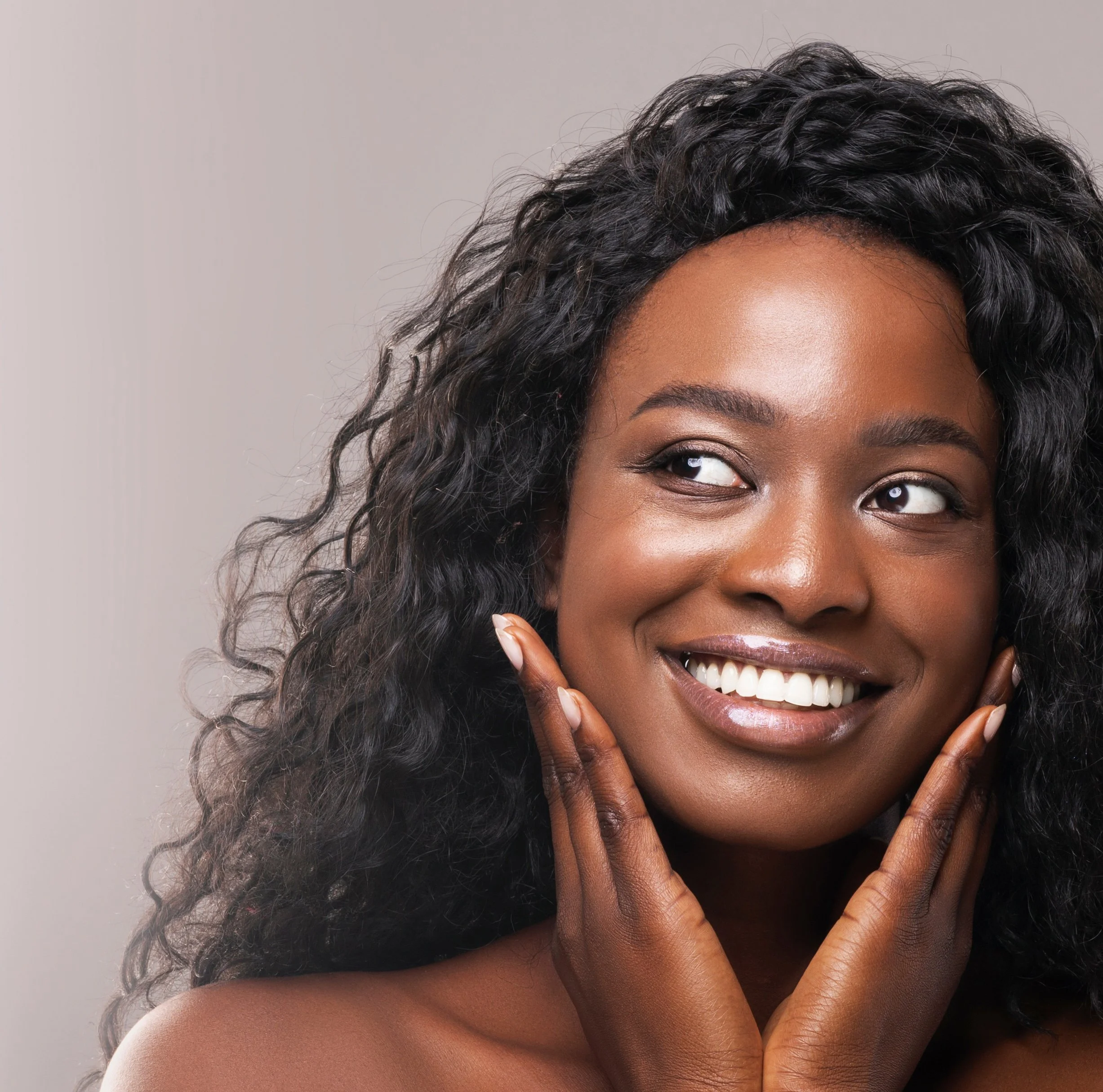 Close-up of a smiling woman with curly black hair and dark skin, touching her face with both hands, against a neutral background.