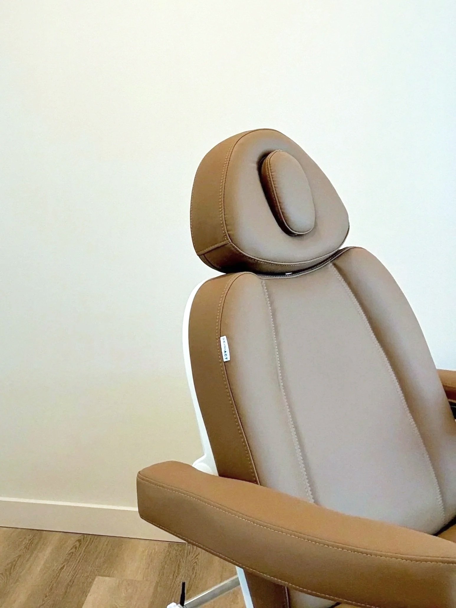 A tan-colored modern office chair with a high backrest and rounded headrest, placed against a plain white wall and hardwood floor.