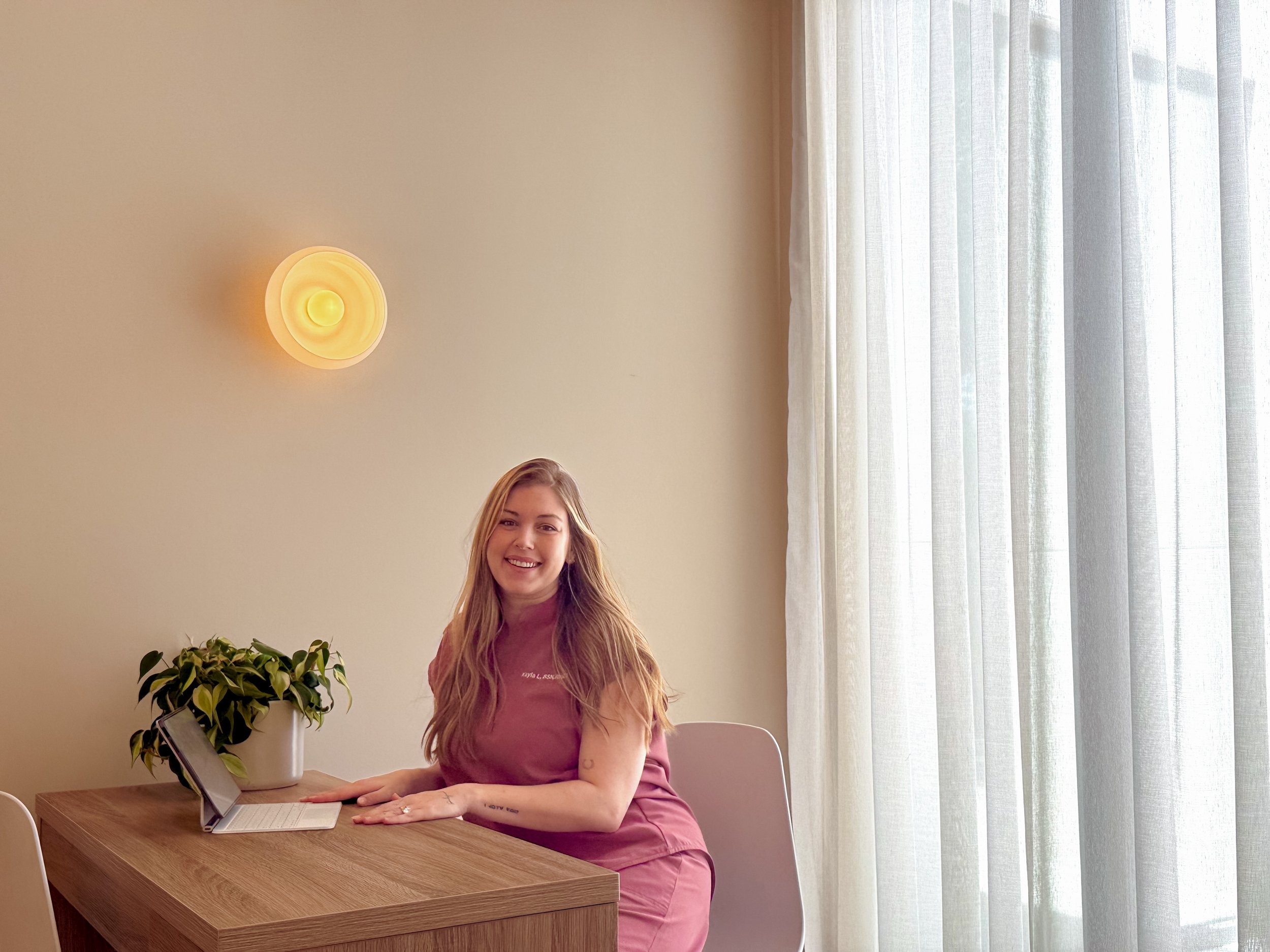 A woman with long reddish hair sitting at a wooden table, smiling, working on a laptop. There is a potted plant on the table, and sheer curtains in the background, along with a wall-mounted yellowish light fixture.