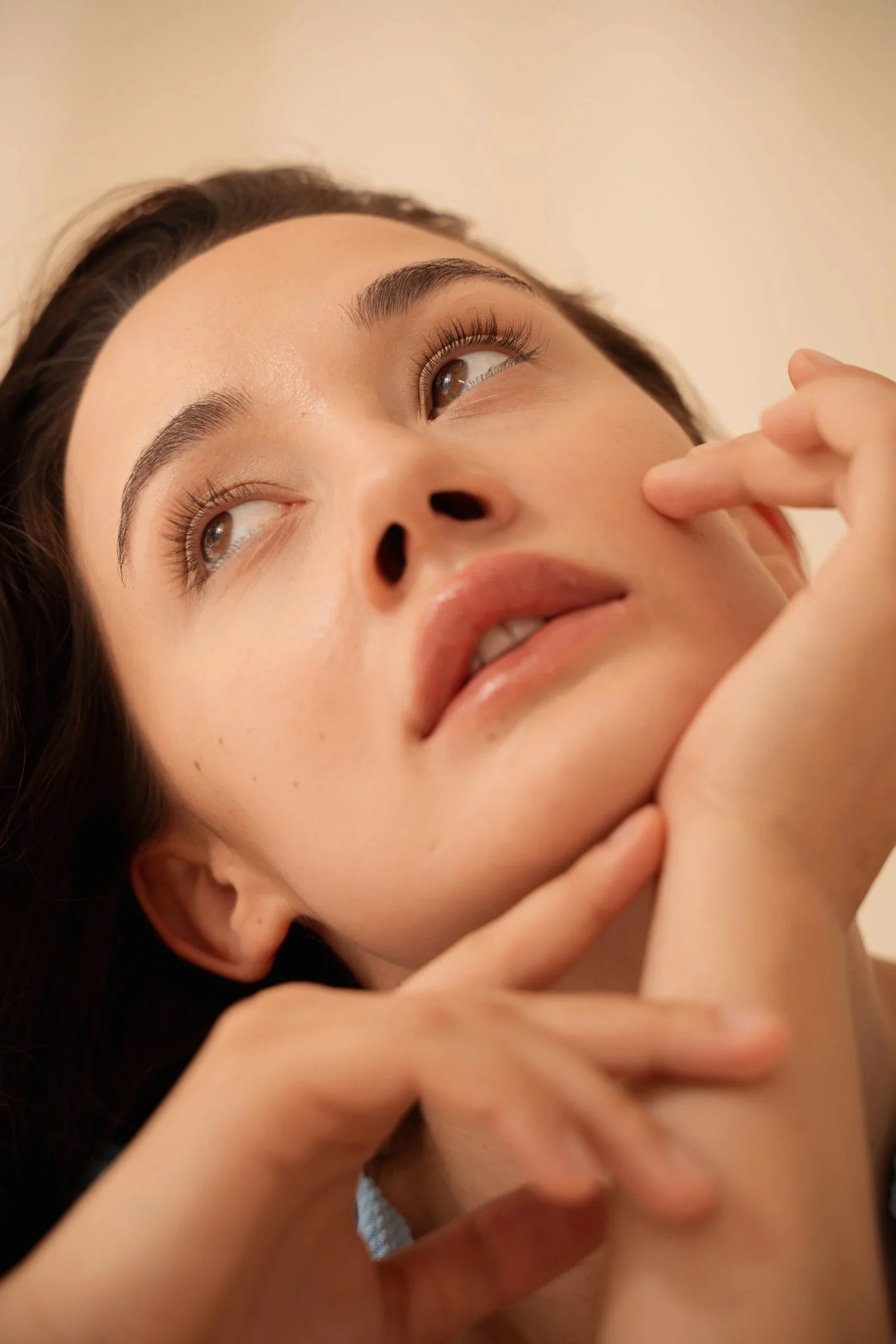 A close-up photo of a woman's face, lying down with her eyes looking upward. She has brown hair, natural makeup, and is touching her cheek with her fingers.