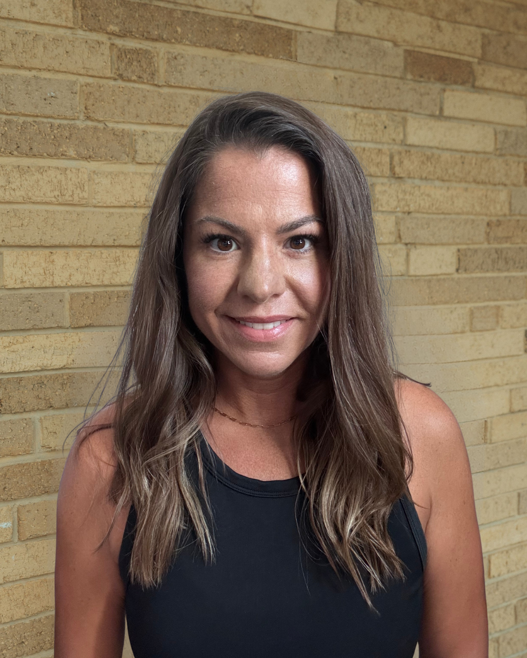 A woman with shoulder-length wavy brown hair, wearing a black sleeveless top and a delicate necklace, smiling in front of a yellow brick wall.