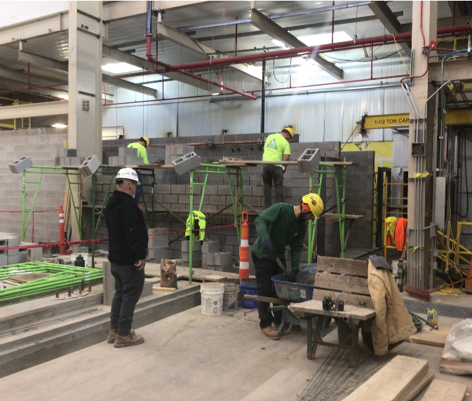 Construction workers building a wall with cinder blocks in an industrial setting, with some workers standing on elevated scaffolding and others working on the ground.