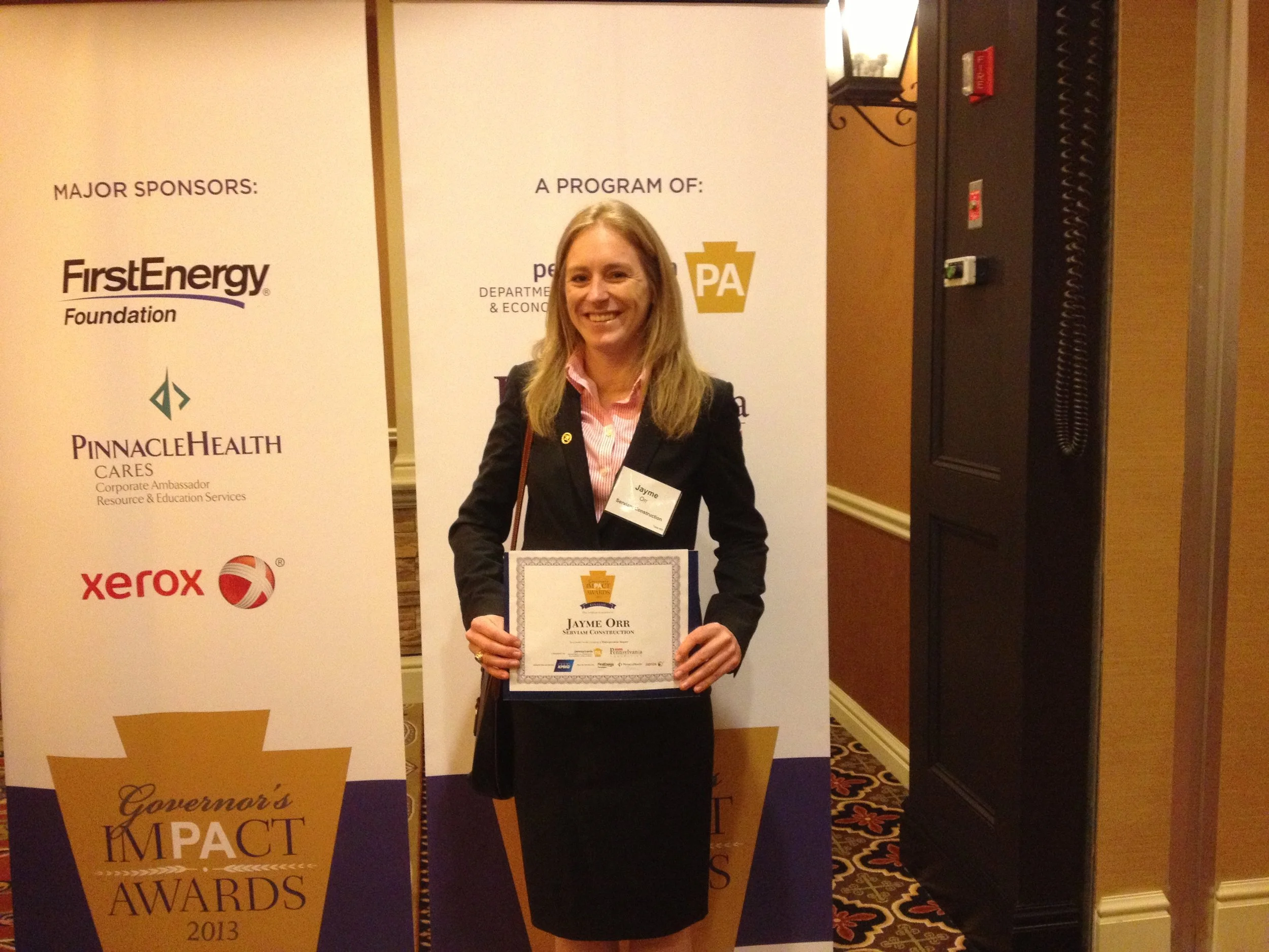 A woman with blonde hair holding a framed certificate, standing in front of a banner with various sponsor logos at an awards event.