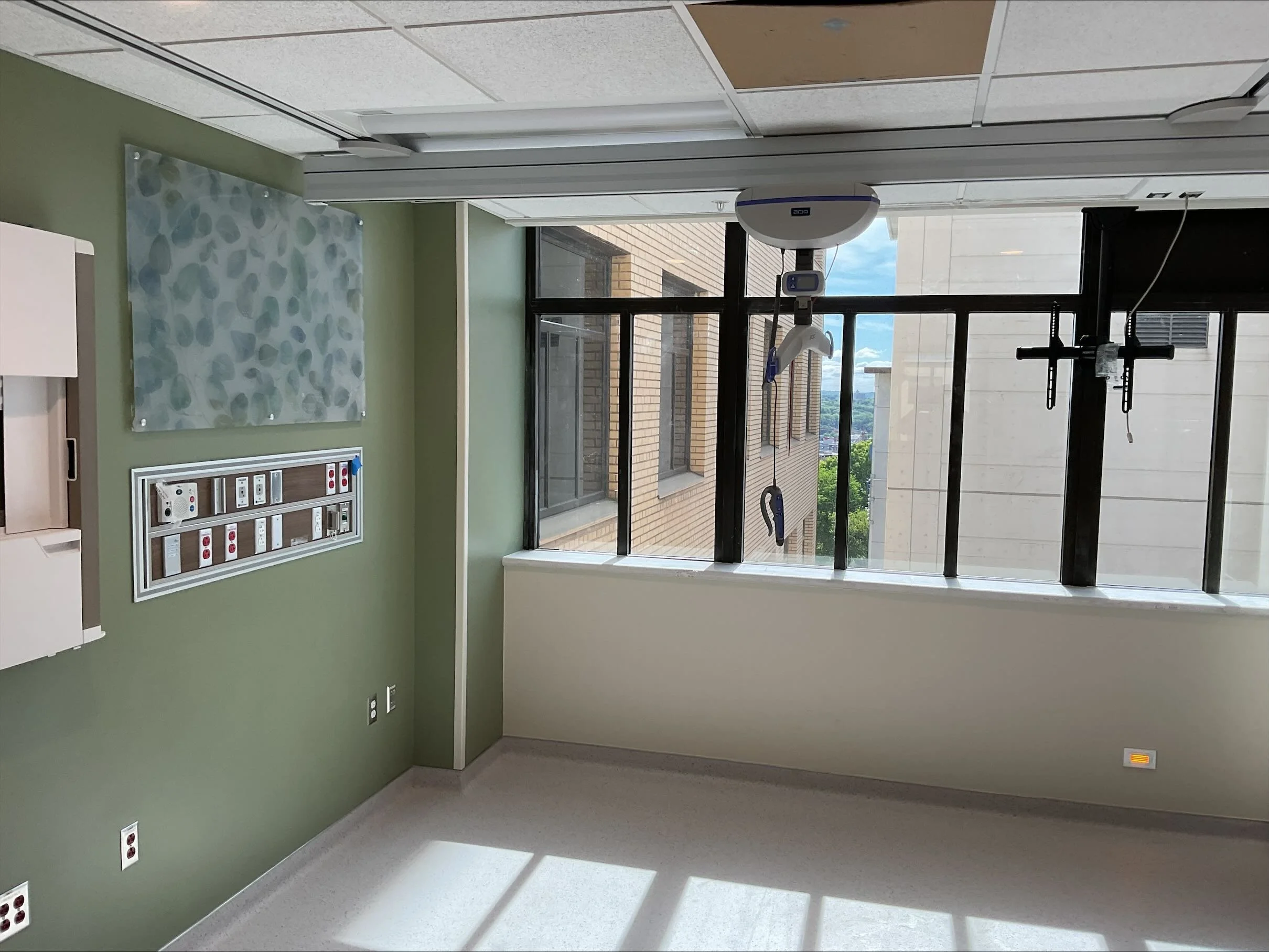 Empty hospital room with green wall, window, and medical equipment, including monitors and medical devices.