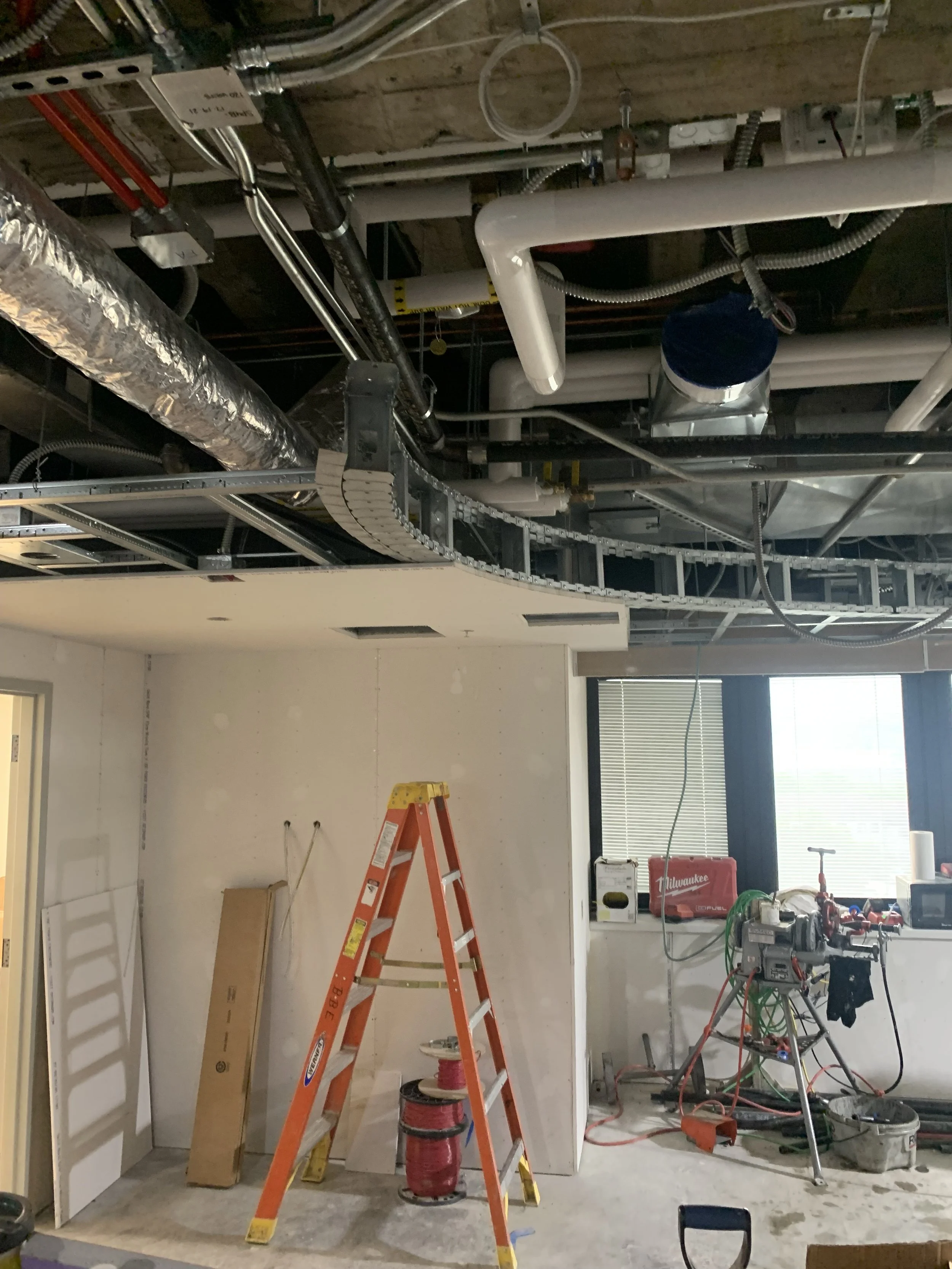 Construction or renovation site with exposed ceiling, pipes, and wiring. An orange ladder stands in the foreground, with various tools and equipment around. There are windows with blinds on the right side.