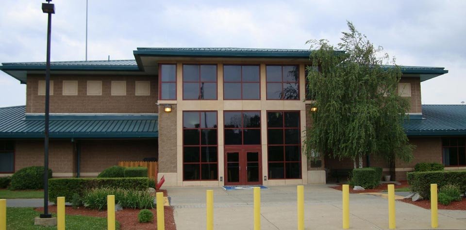Exterior of a modern two-story building with large glass windows, a green metal roof, landscaped bushes, and a tree, with yellow posts at the entrance.