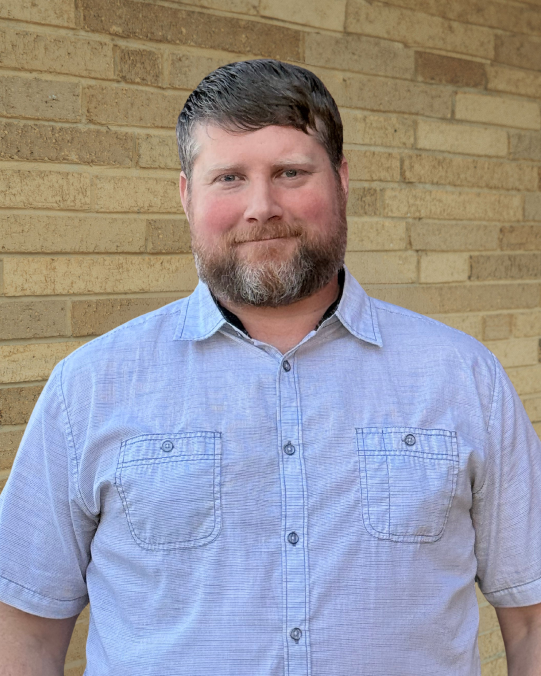 A man with a beard and short hair wearing a light gray collared shirt, standing in front of a brick wall.
