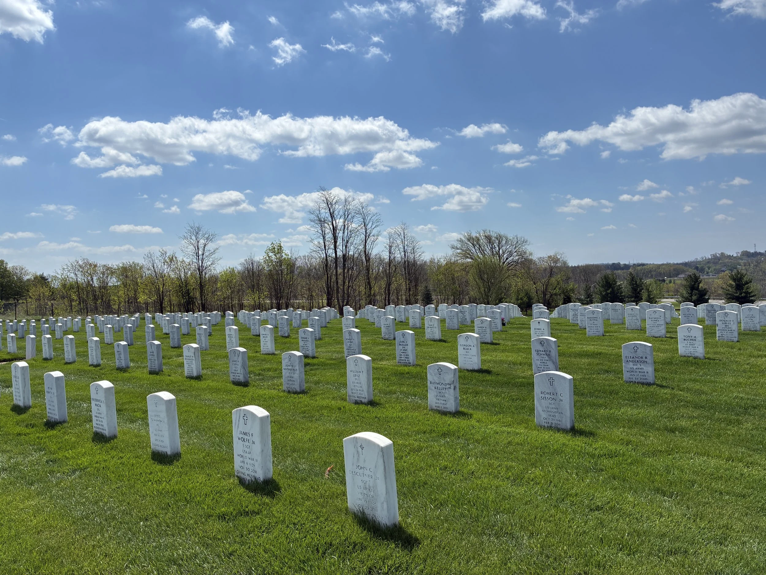A large cemetery with rows of white gravestones in a green field under a bright blue sky with scattered clouds. Trees are visible in the background.