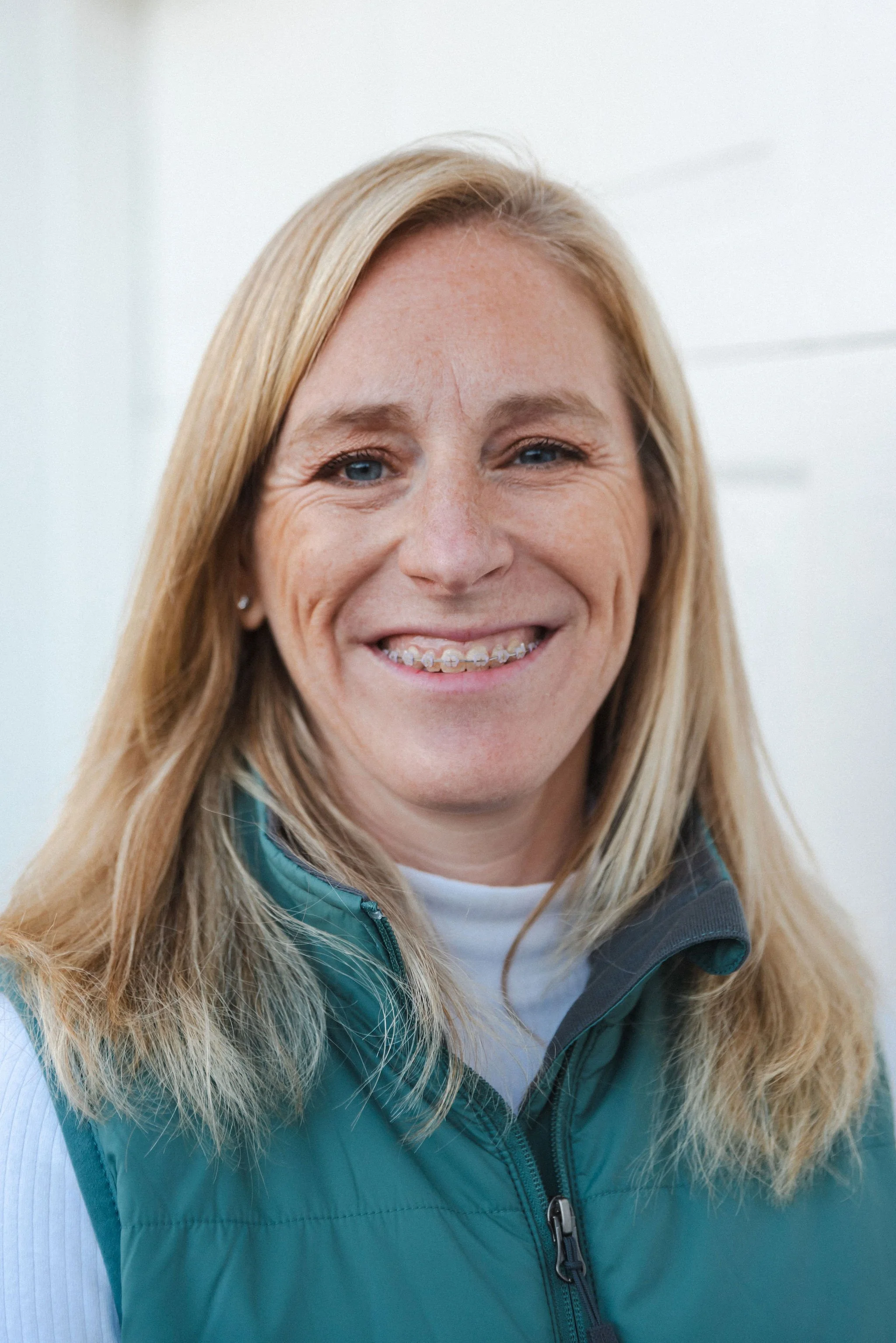 Close-up of a smiling woman with blonde hair, blue eyes, braces, wearing a teal jacket and white shirt, standing indoors with a white background.
