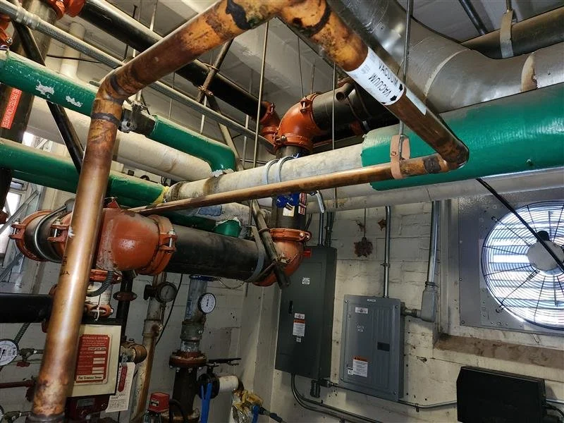 Indoor industrial mechanical room with pipes, valves, gauges, and electrical panels.
