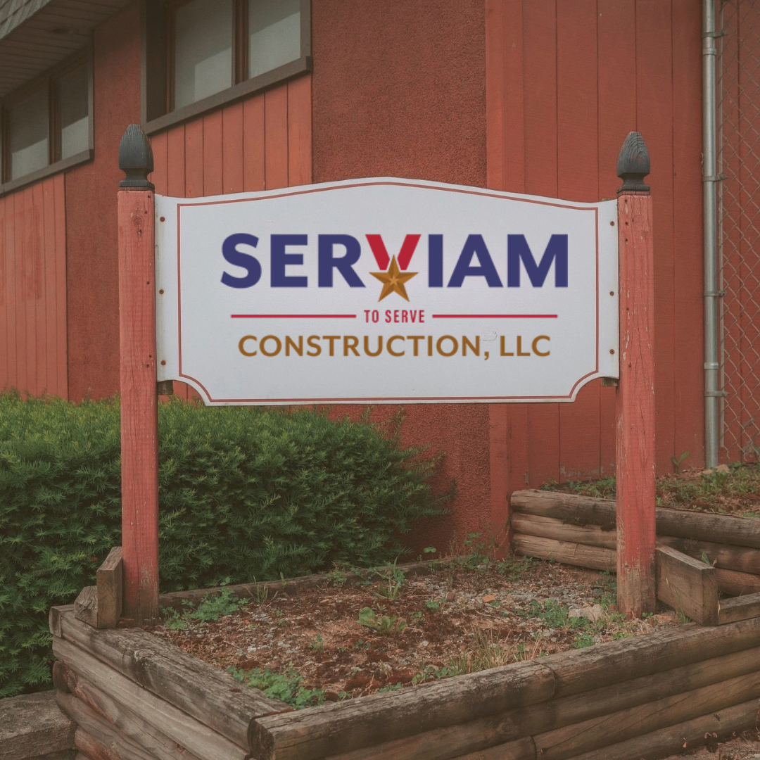 A sign for Serviam Construction, LLC, with patriotic design elements such as red, white, and blue colors and a star, mounted on red wooden posts outside a building with red siding and a chain-link fence to the side.