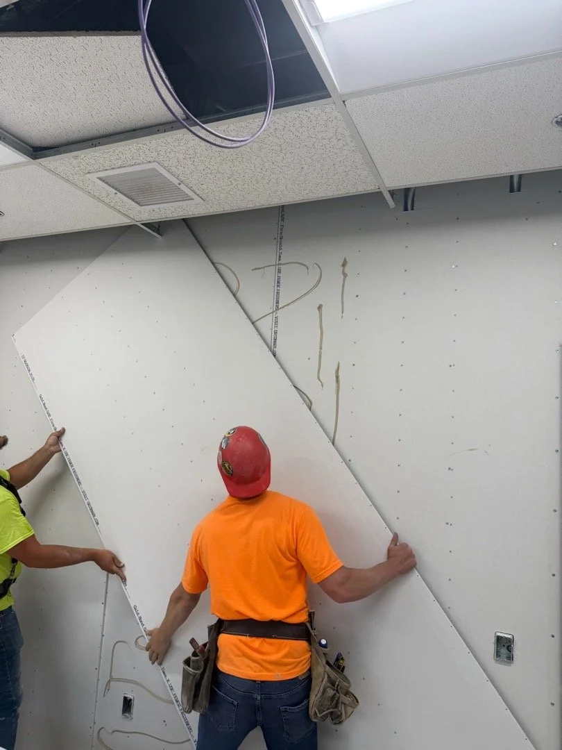 Two construction workers installing drywall on a wall in an office space, with part of the ceiling and electrical wiring visible.