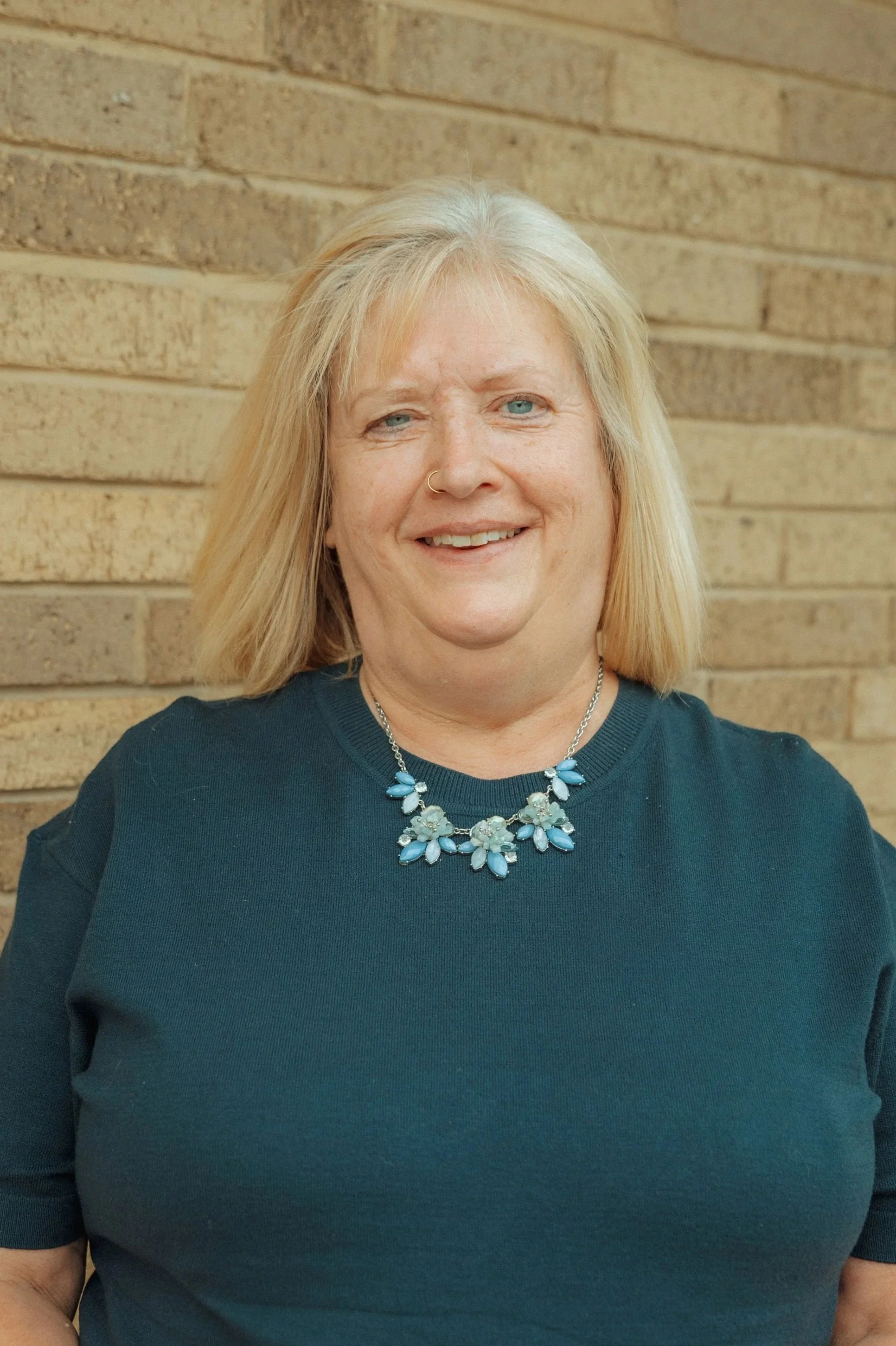 Close-up of a woman with blonde hair, blue eyes, wearing a dark green top and a statement necklace with blue and clear stones, standing against a brick wall.