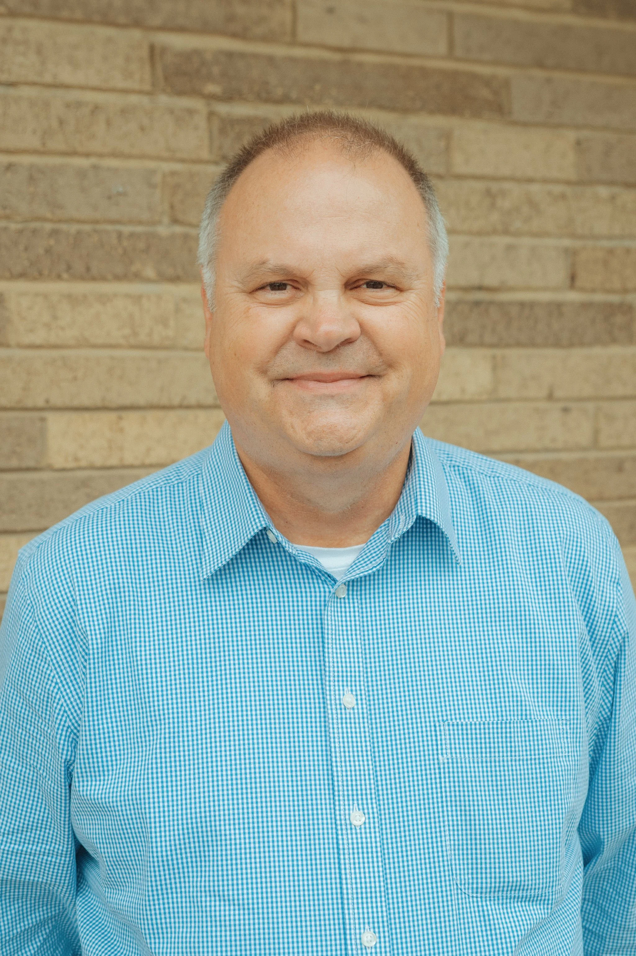 A headshot of a middle-aged man with light skin, short hair, and a slight smile, wearing a blue checked shirt, standing in front of a brick wall.
