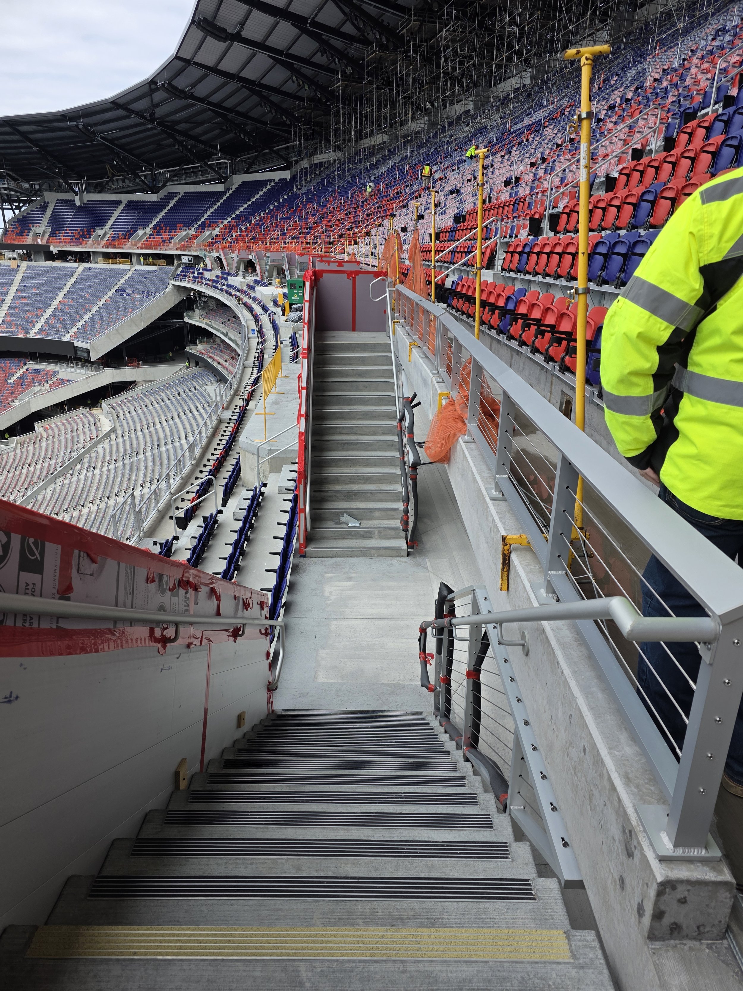 Construction workers install seats on the upper tiers of a stadium. The seats are red and blue, and some are covered with orange safety netting. There are stairways with yellow tactile paving, and a person in a yellow safety vest is partially visible on the right.