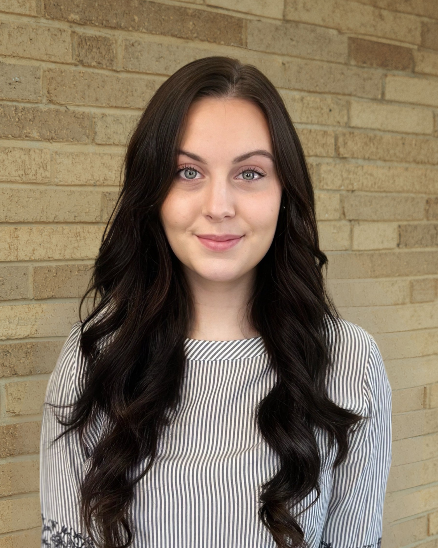 A young woman with long wavy dark hair and light skin smiling in front of a brick wall, wearing a striped shirt.