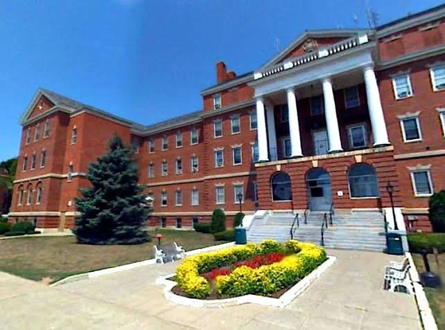 Red brick building with white columns and steps leading up to the entrance, landscaped flower bed in front with yellow and red flowers, trees, blue sky.