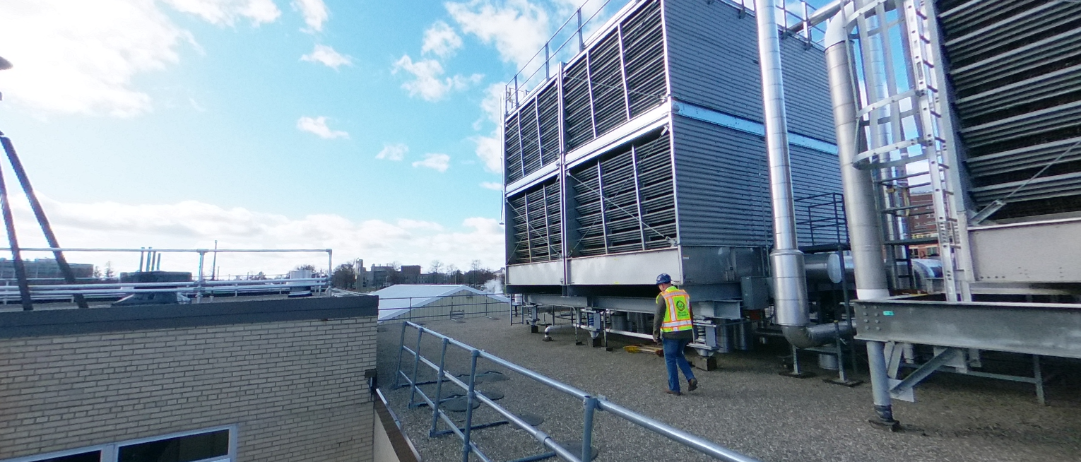 A worker in a neon safety vest and helmet walks near large HVAC units on a building rooftop during daytime.