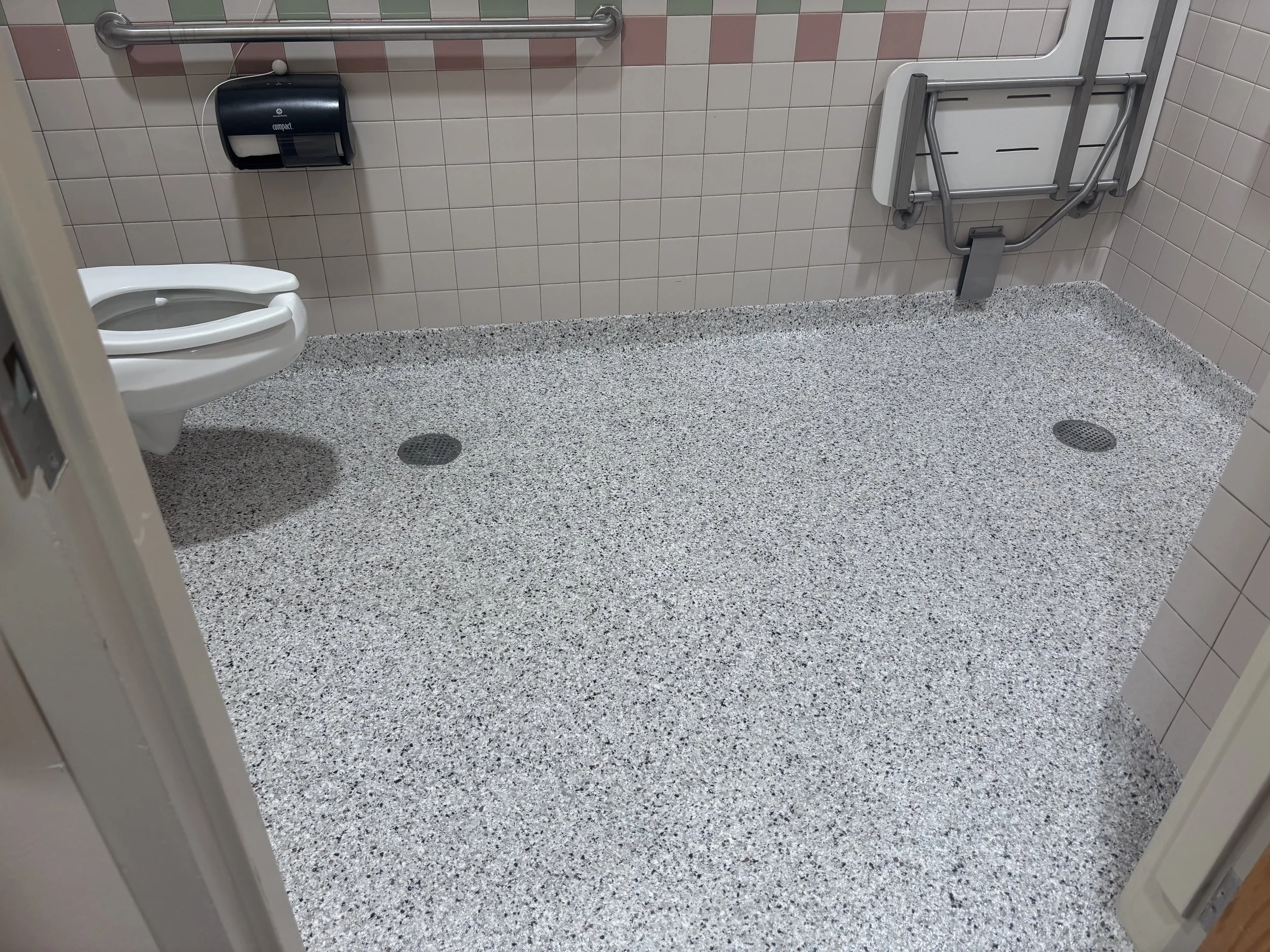 A bathroom with pink and white tiled walls, terrazzo flooring, a white toilet, a black paper towel dispenser, and a wall-mounted foldable shower chair.