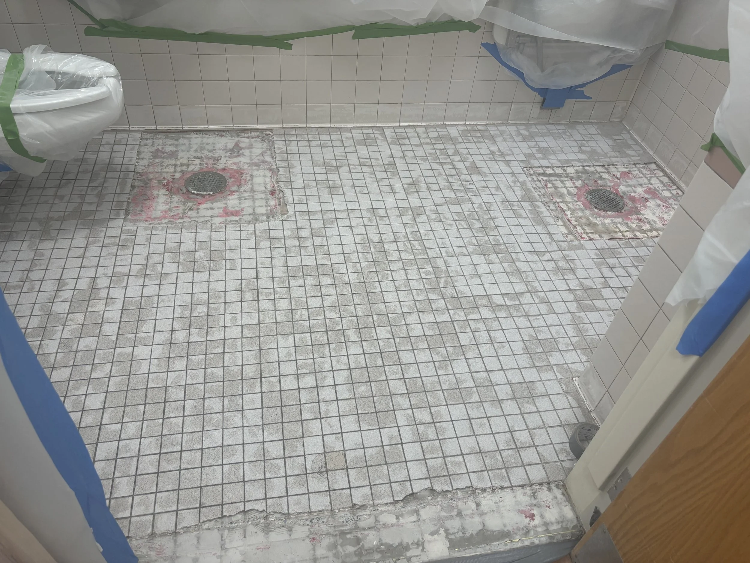 Bathroom with tile floor and wall, two floor drain covers, and areas under renovation with missing tiles and exposed adhesive, partially covered by plastic sheeting and painter's tape.
