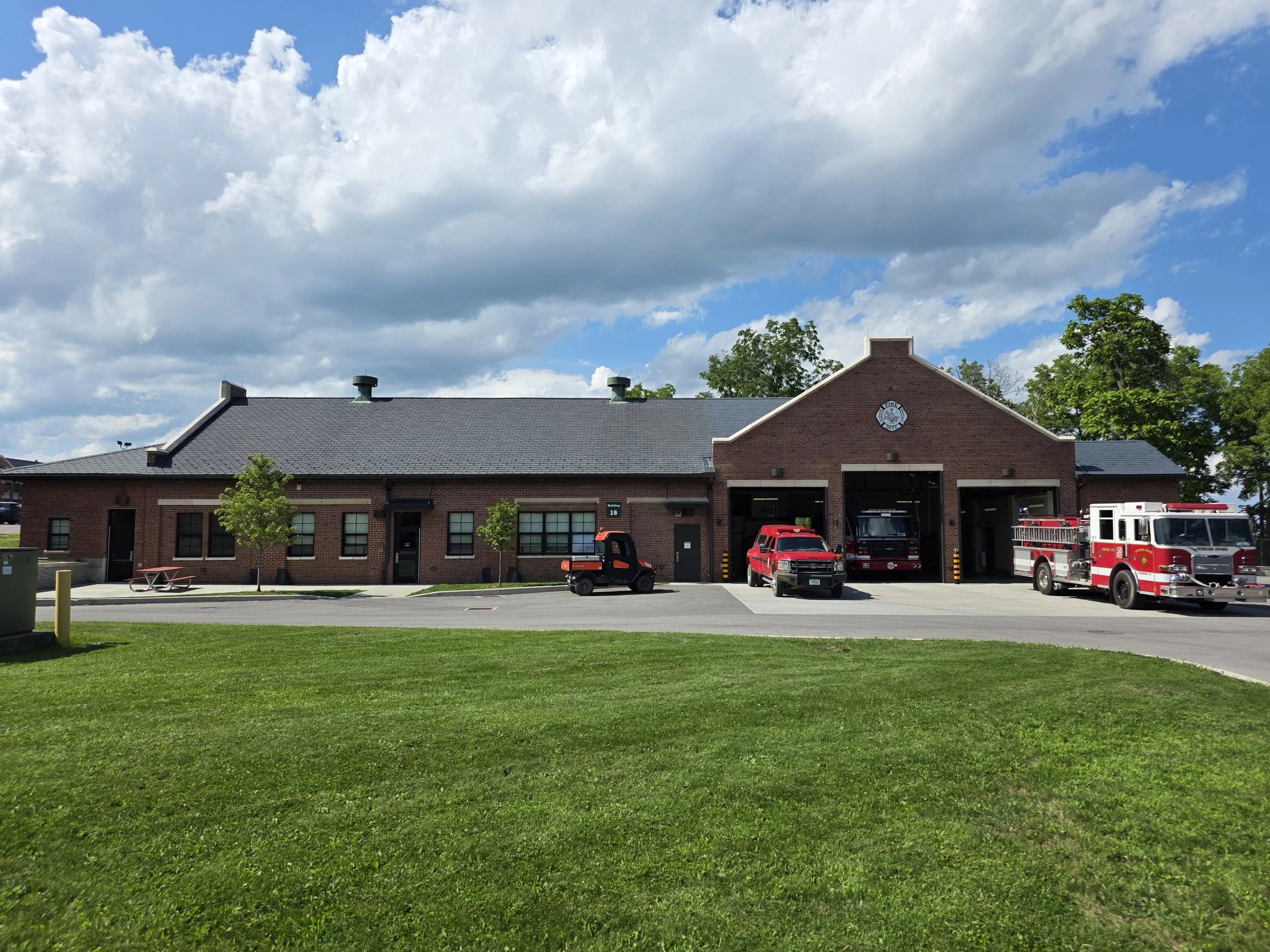 A fire station building with a brick facade and a fire department emblem on the front, surrounded by a grassy lawn, with parked fire trucks outside and a partly cloudy sky overhead.