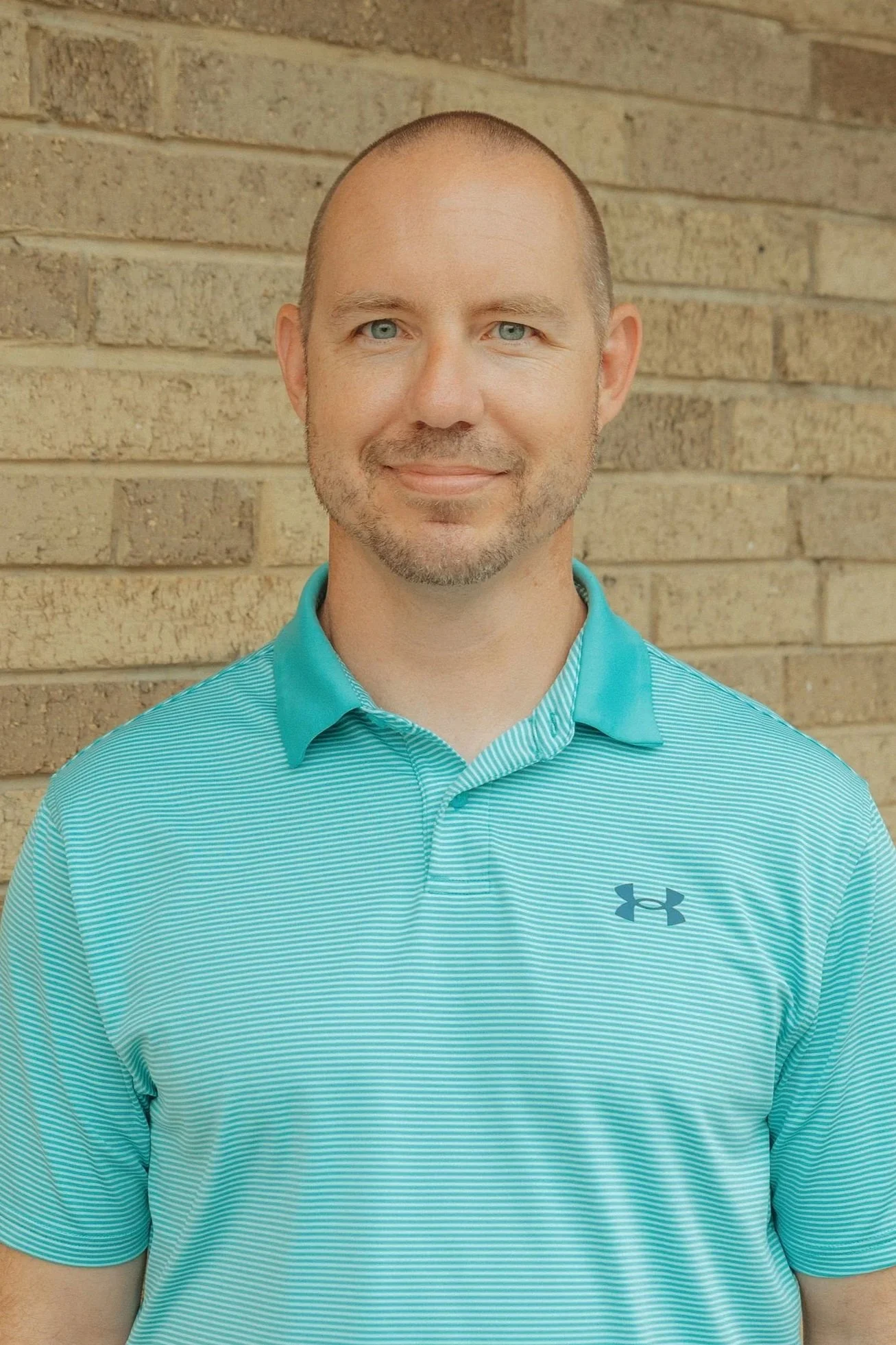 Portrait of a smiling man with light skin, blue eyes, and a shaved head, wearing a teal Under Armour polo shirt, standing against a brick wall background.