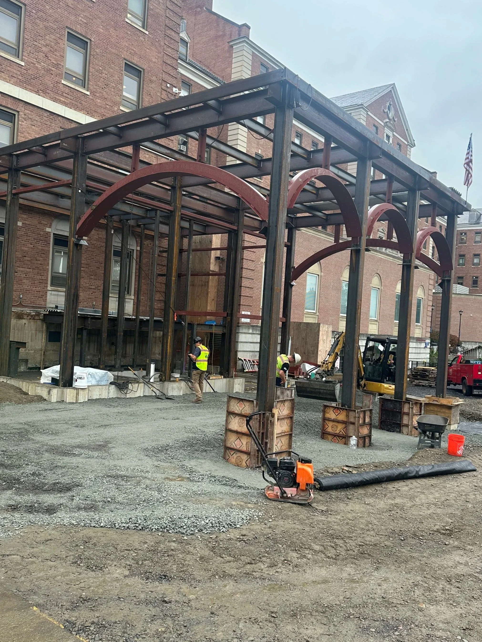 Construction site with steel framework and workers in yellow safety vests and helmets. Building walls in background, construction equipment, and scattered materials on ground.
