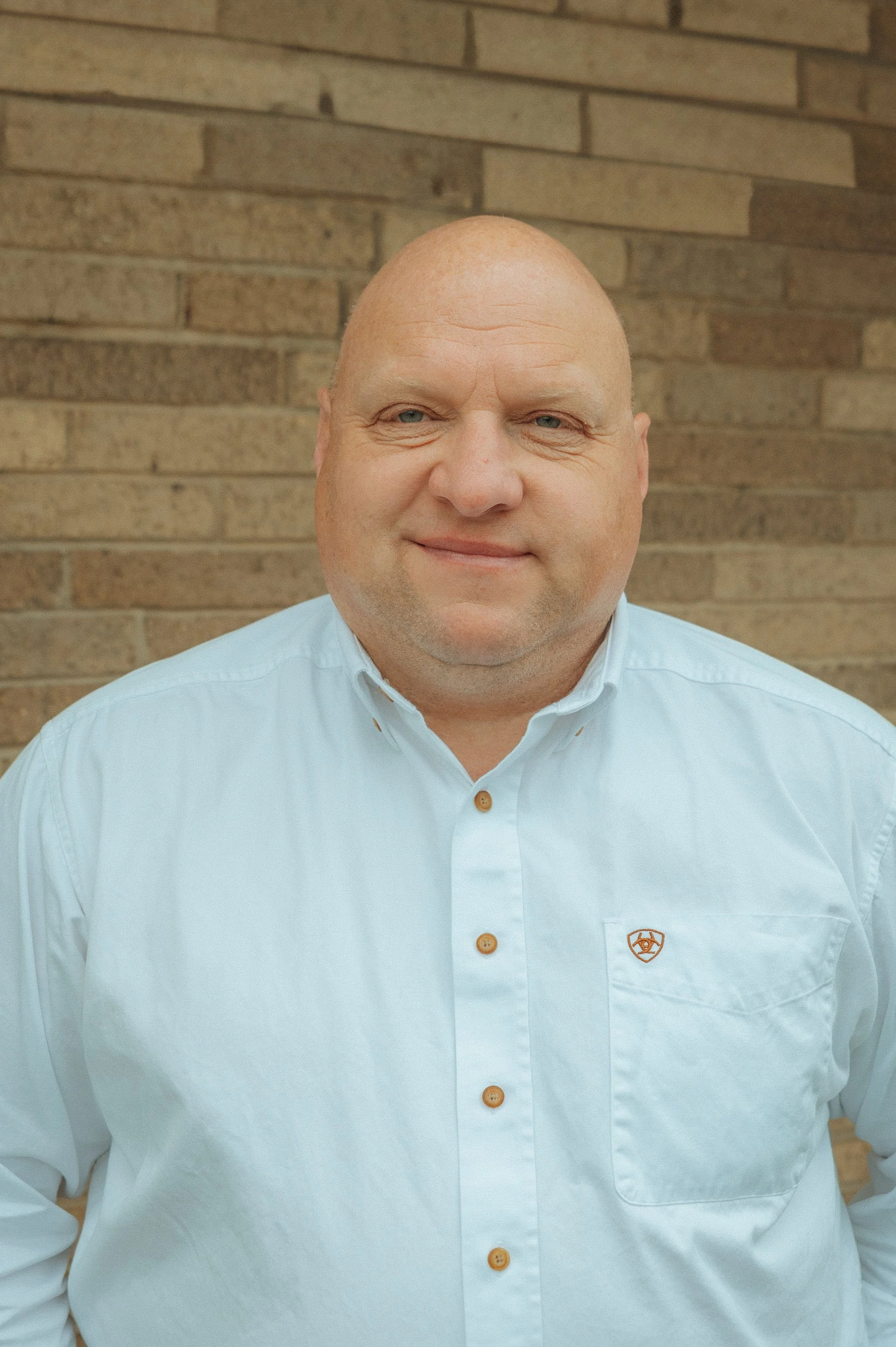 Close-up of a bald man wearing a white shirt with a logo, standing in front of a brick wall.