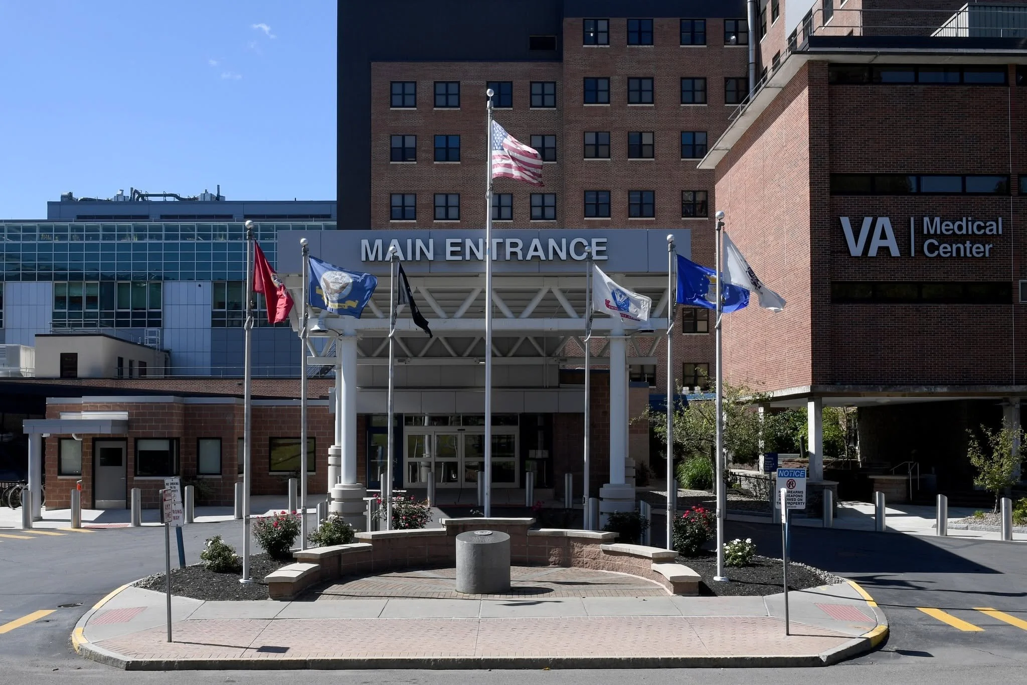 Medical center building with a sign that reads 'Main Entrance' and flags flying in front. The building is made of brick and glass, with multiple windows. The sky is clear and blue.
