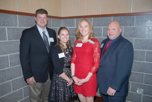 Four people standing together indoors against a brick wall, dressed in formal attire, smiling at the camera.