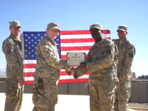 Four soldiers in uniform standing in front of an American flag, exchanging a box or award.